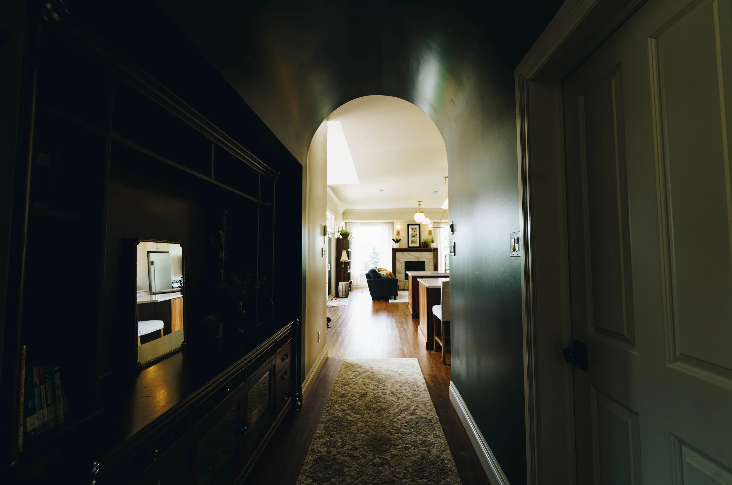 Hallway leading to a living room with a fireplace, armchair, and large window; dark bookshelf on the left and a door on the right.