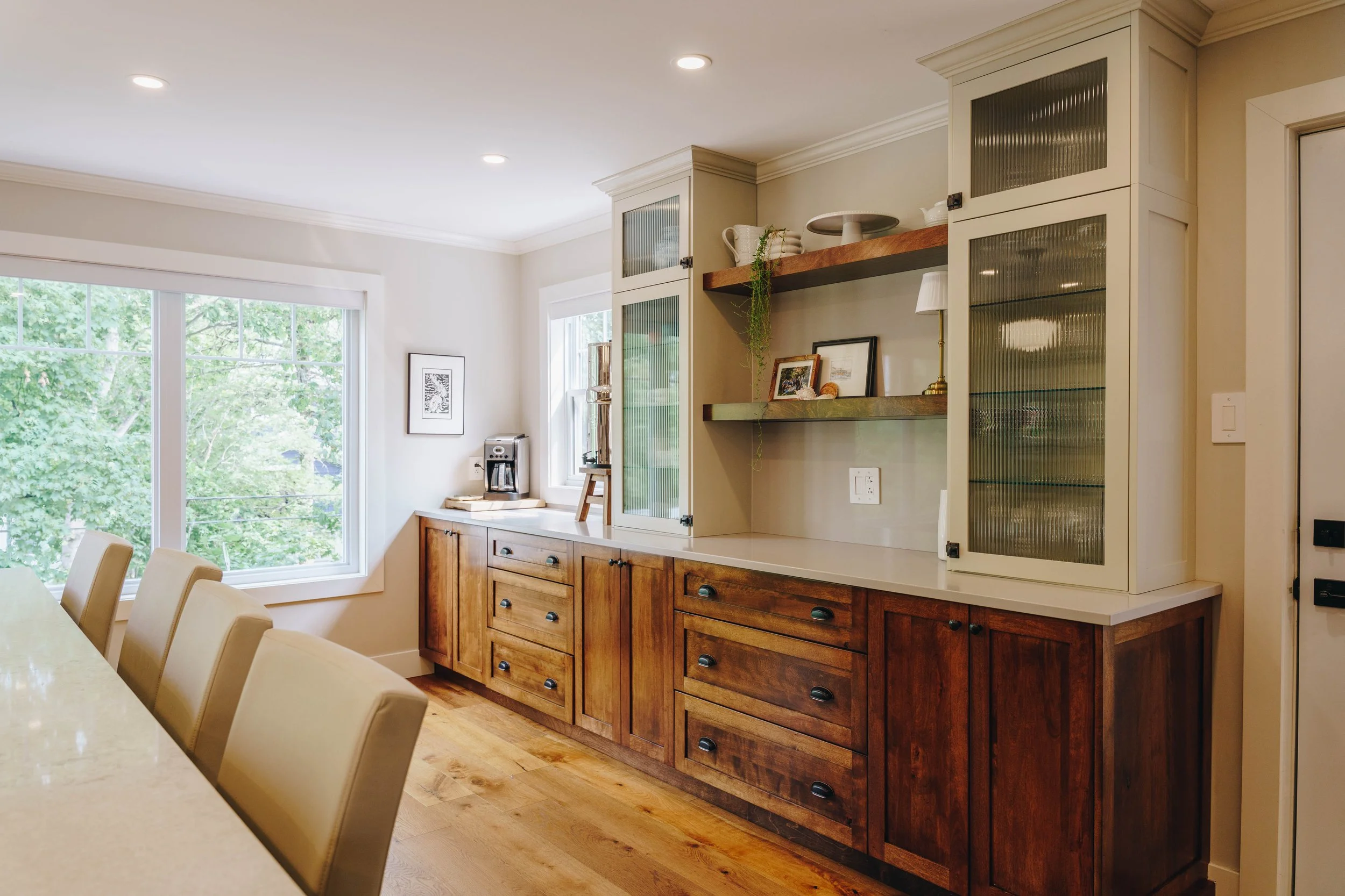 Kitchen with wooden cabinets, open shelving, a coffee maker on the counter, and large windows showing green trees outside.