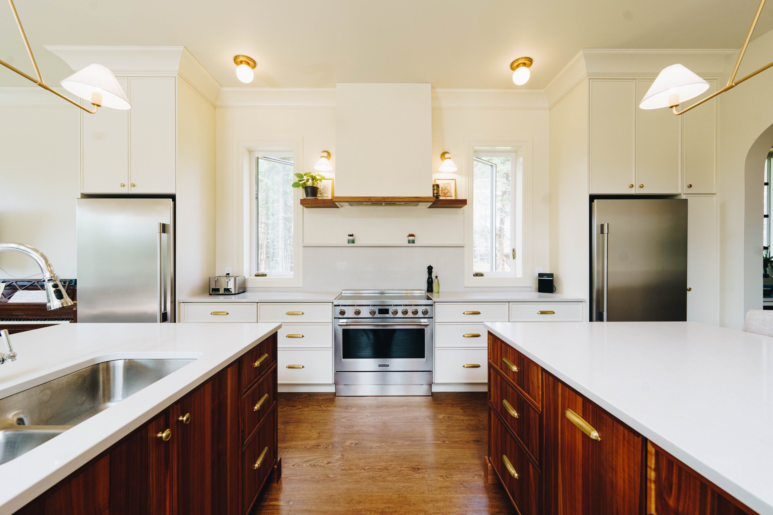 Modern kitchen with white cabinets, stainless steel appliances, and wooden island with gold handles, featuring pendant and ceiling lights, windows behind stove, and hardwood floors.