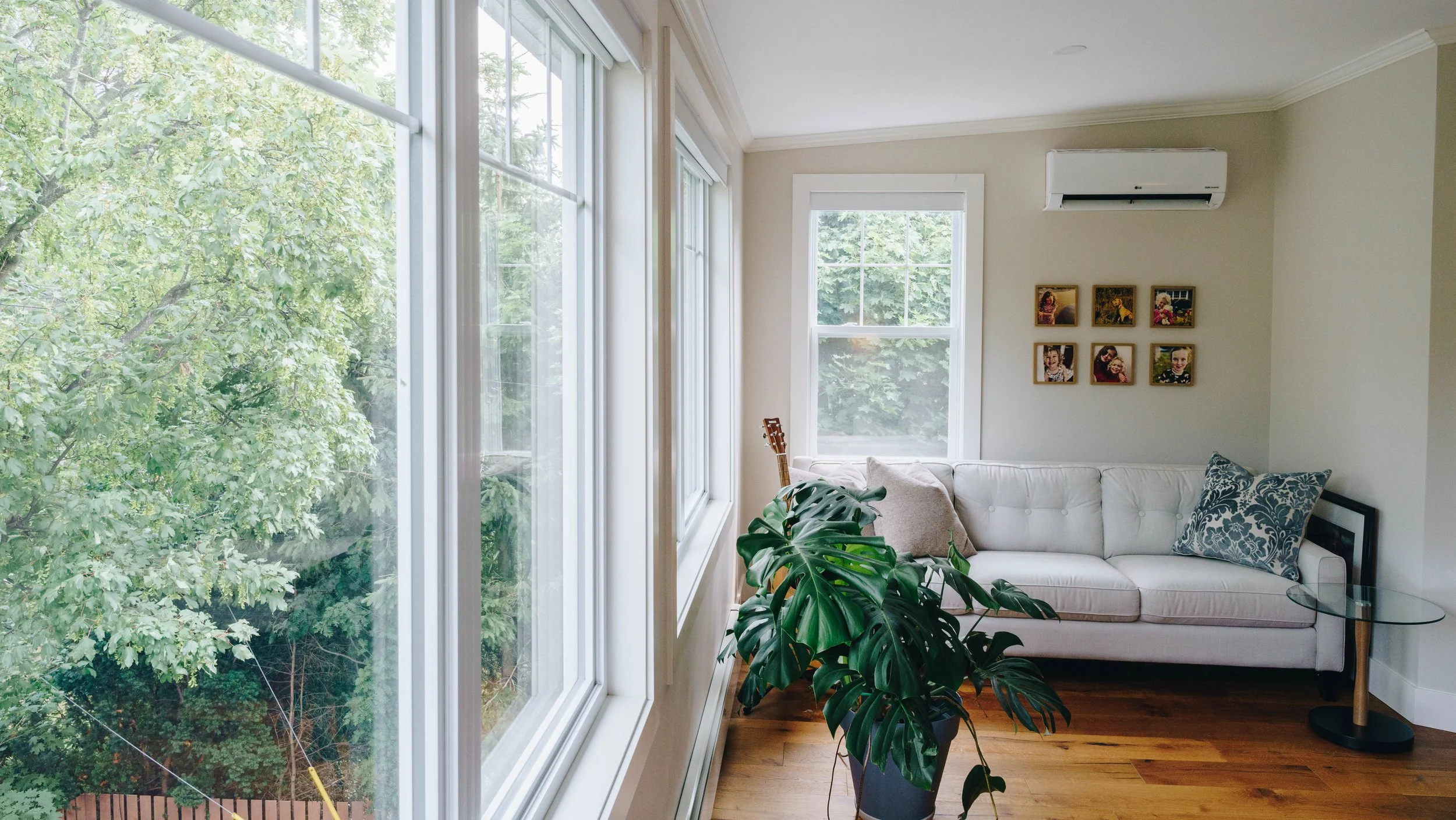 Living room with large windows, white sofa, decorative pillows, framed photos on the wall, houseplants, and hardwood floors.