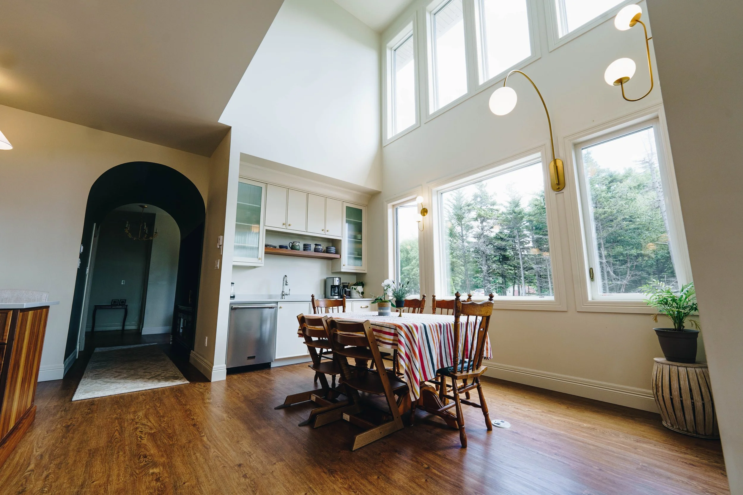 Bright, modern dining area with large windows, a wooden table with a colorful striped tablecloth, and wooden chairs. White kitchen cabinets and greenery outside visible.