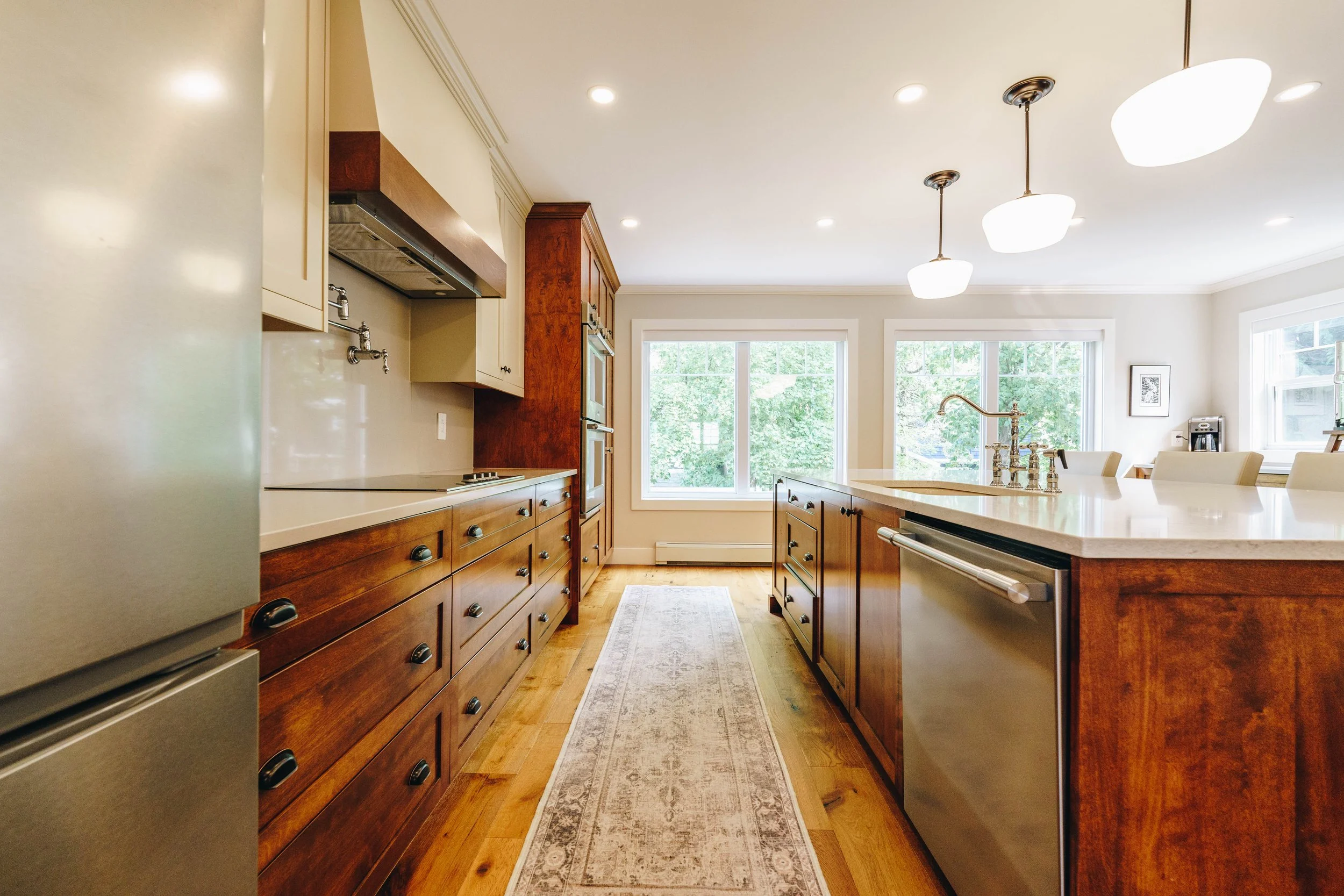 Bright kitchen with wooden cabinets, a large island with a sink, and breakfast bar chairs, illuminated by hanging lights and natural light from large windows.