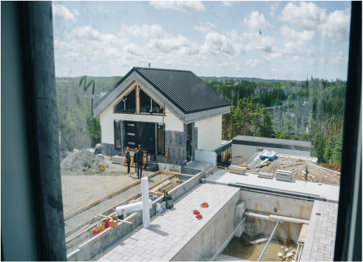 Construction site with a house being built, workers present, surrounded by construction materials and equipment, overlooking a wooded landscape.