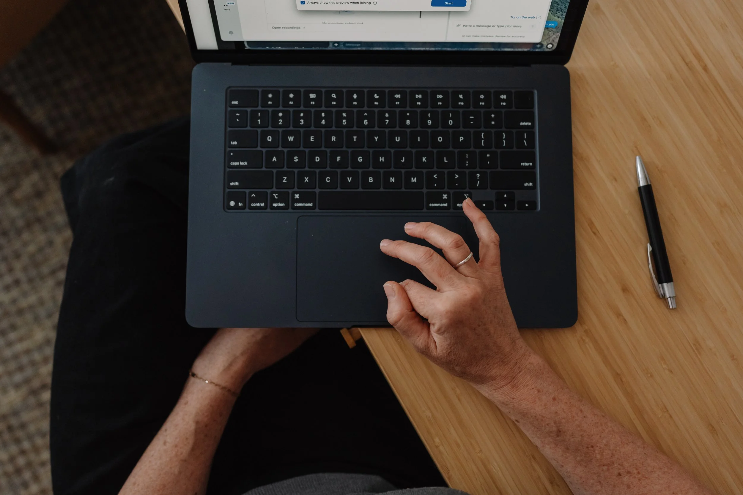 A person using a laptop with a black keyboard on a wooden table, with a black pen lying nearby. The person is touching the keyboard with one hand.