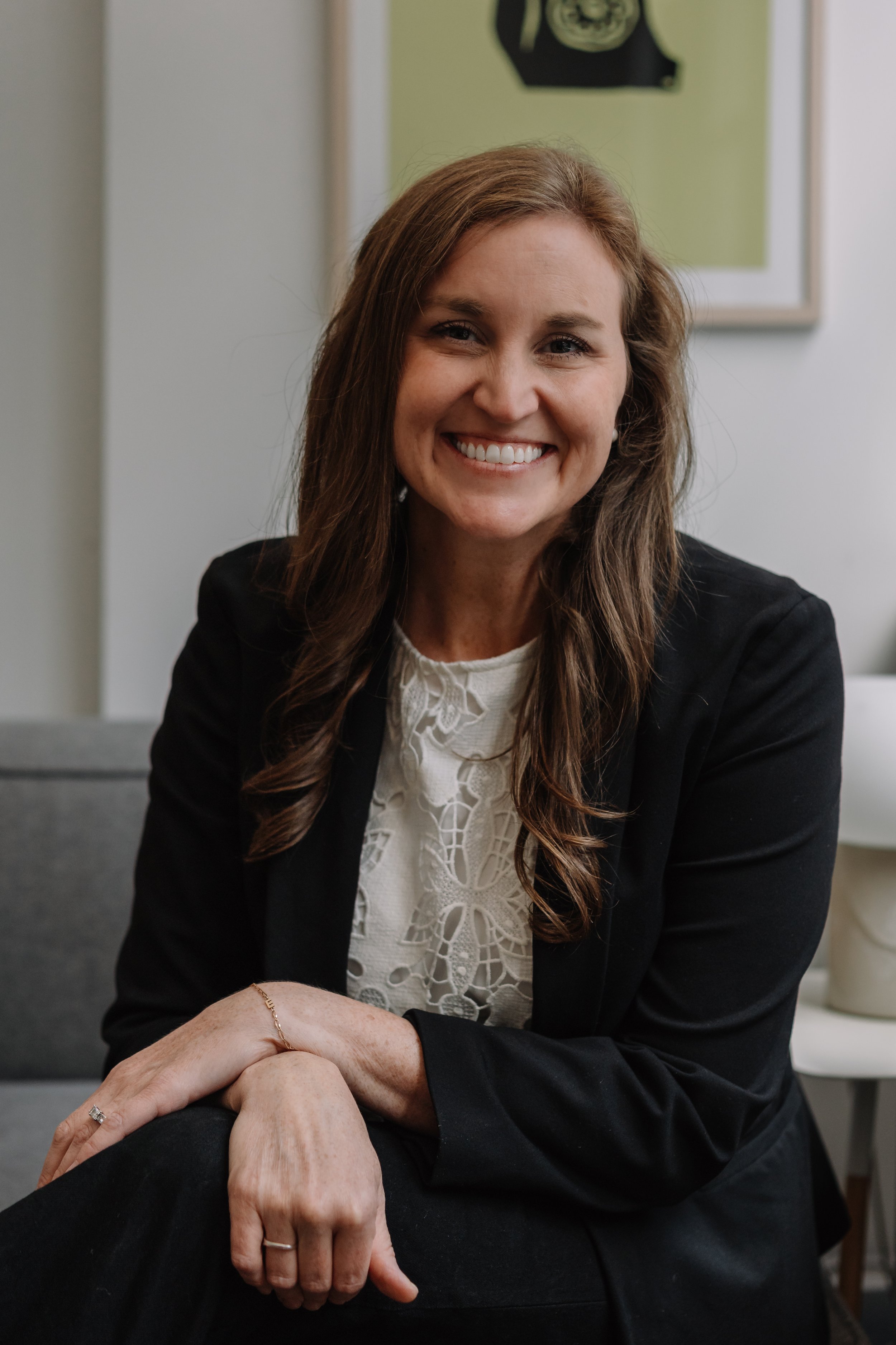 A woman with long, wavy brown hair and a big smile, wearing a black blazer over a white lace top, sitting indoors with a framed picture on the wall in the background.