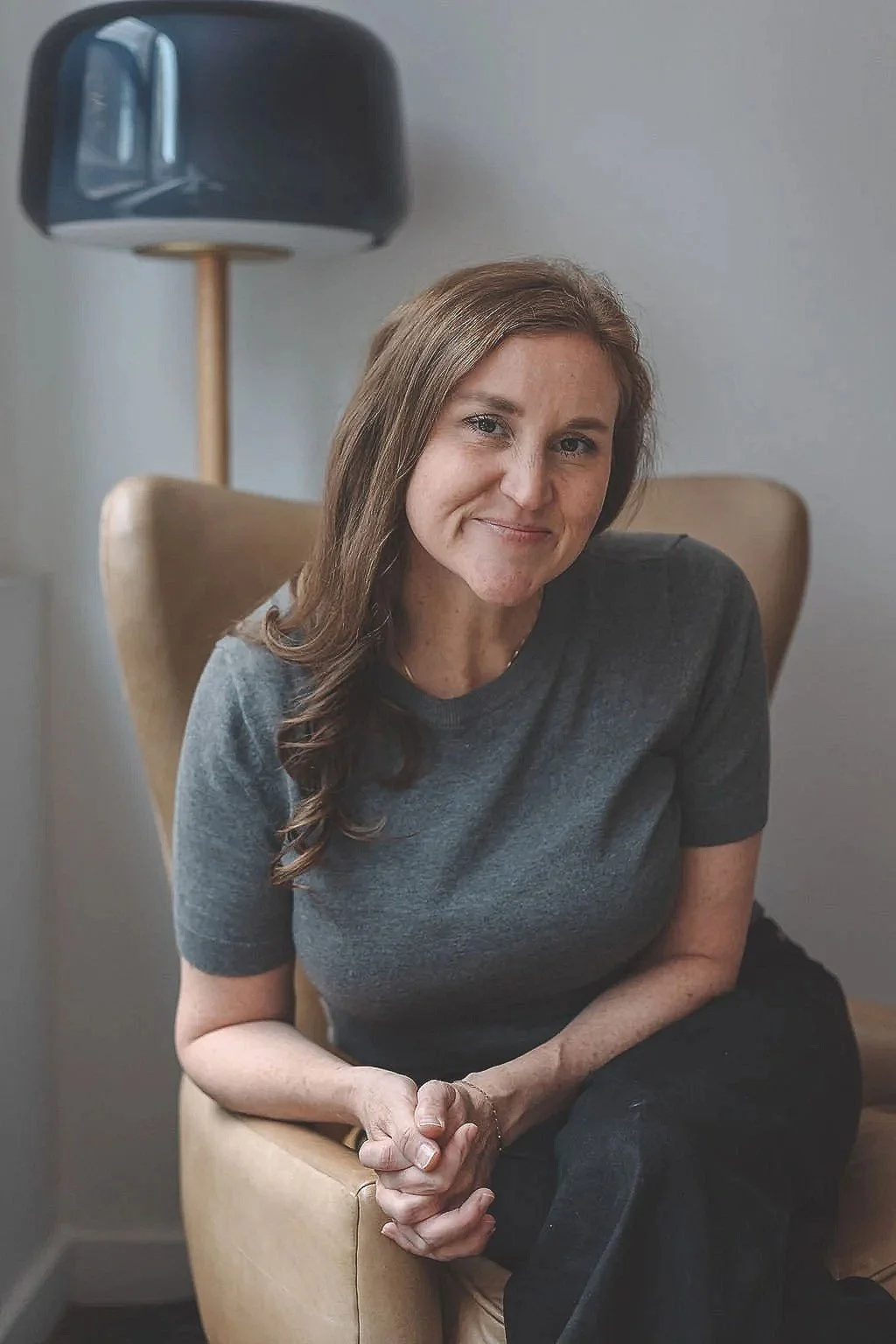 A woman with long brown hair sitting in a beige armchair, smiling at the camera, wearing a dark gray t-shirt, with a black lamp and a plain wall in the background.