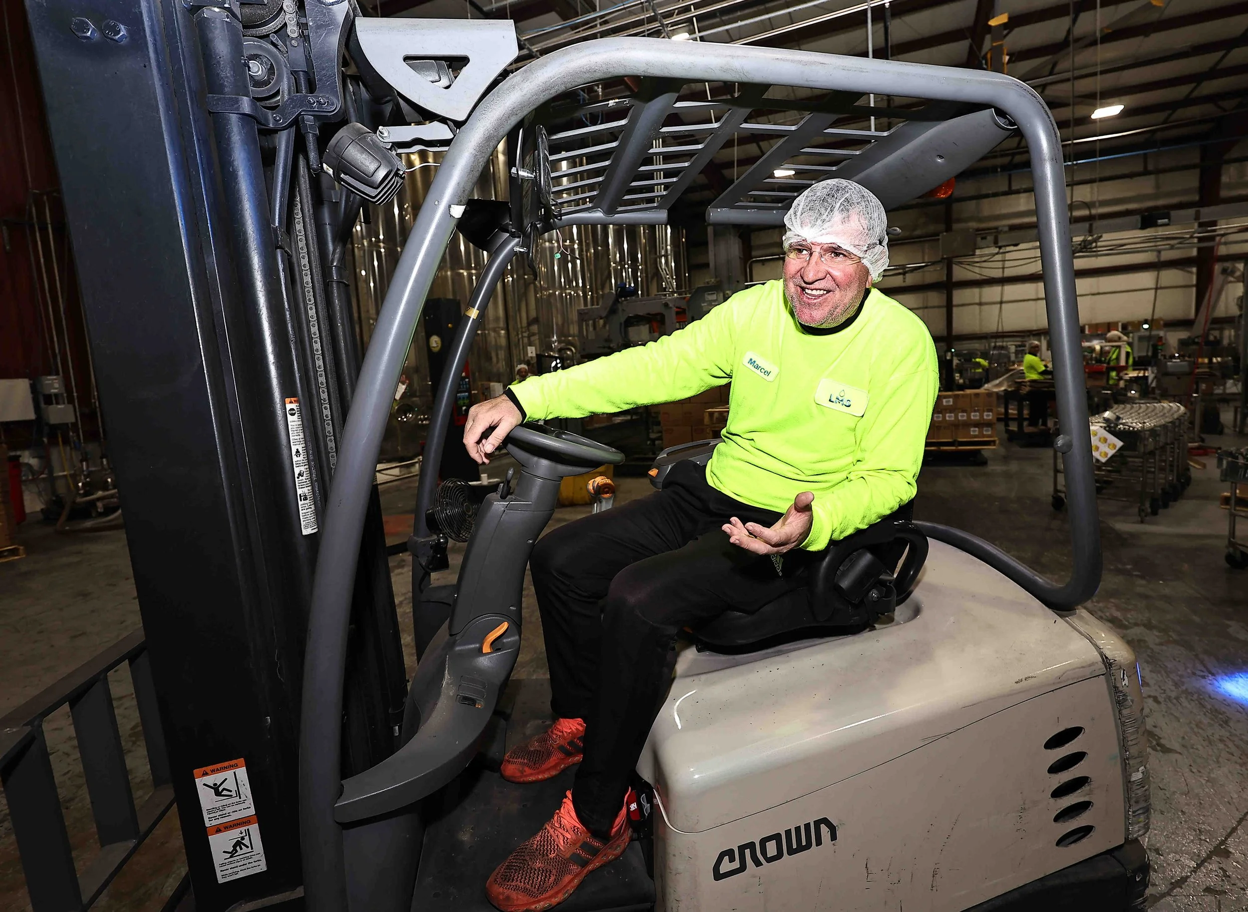 A male employee sitting on a forklift inside a warehouse, wearing a yellow shirt, a hairnet, and red sneakers, smiling and gesturing with his hand.