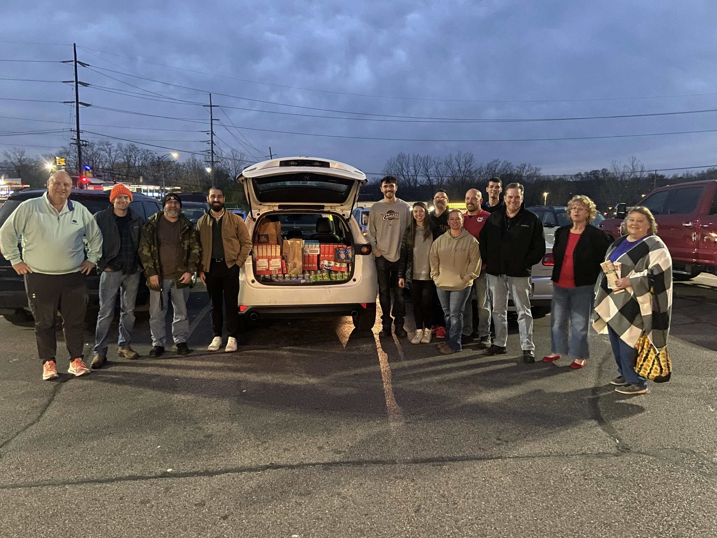 A group of employees standing in a parking lot around an open trunk filled with food and supplies, for a food drive.