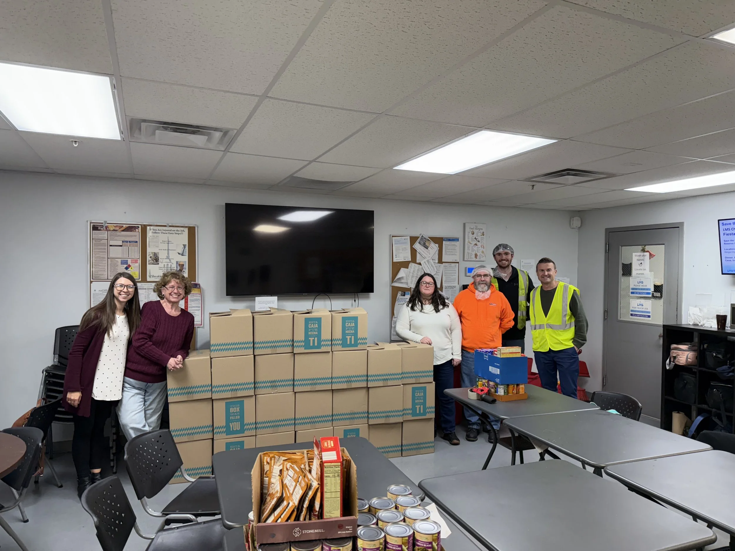 Group of six people standing in a room with donation boxes and food items, smiling for the camera.