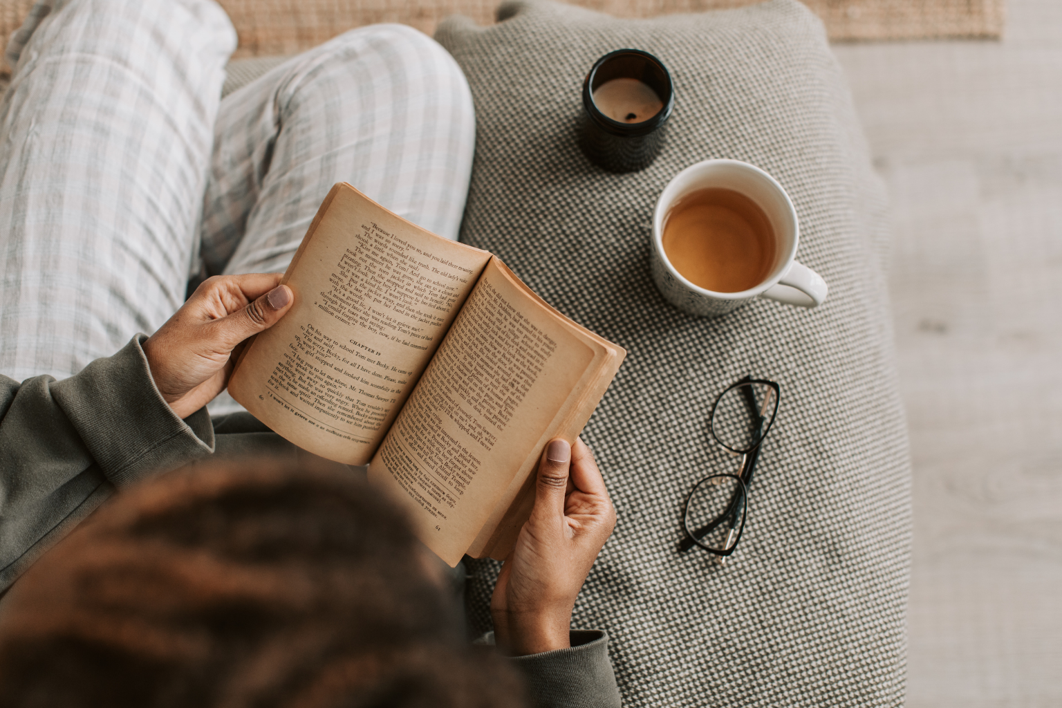 Person sitting on a couch with striped pajamas, reading a paperback book, with a mug of tea, a black candle, and glasses on a textured table nearby.