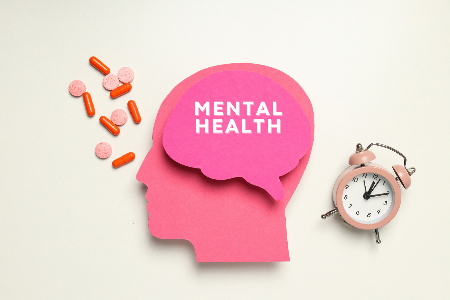 Pink paper cutout of a human head with a speech bubble containing 'MENTAL HEALTH', scattered pills, and a pink alarm clock on a white background.