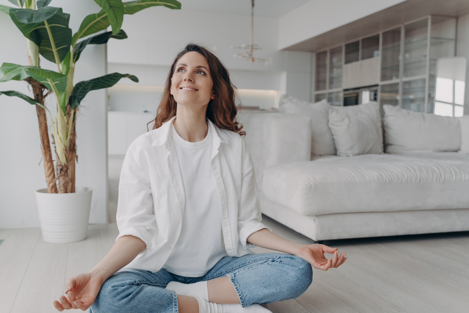 A woman practicing yoga and meditation while sitting cross-legged on the floor in a bright, modern living room with a large potted plant and white furniture.