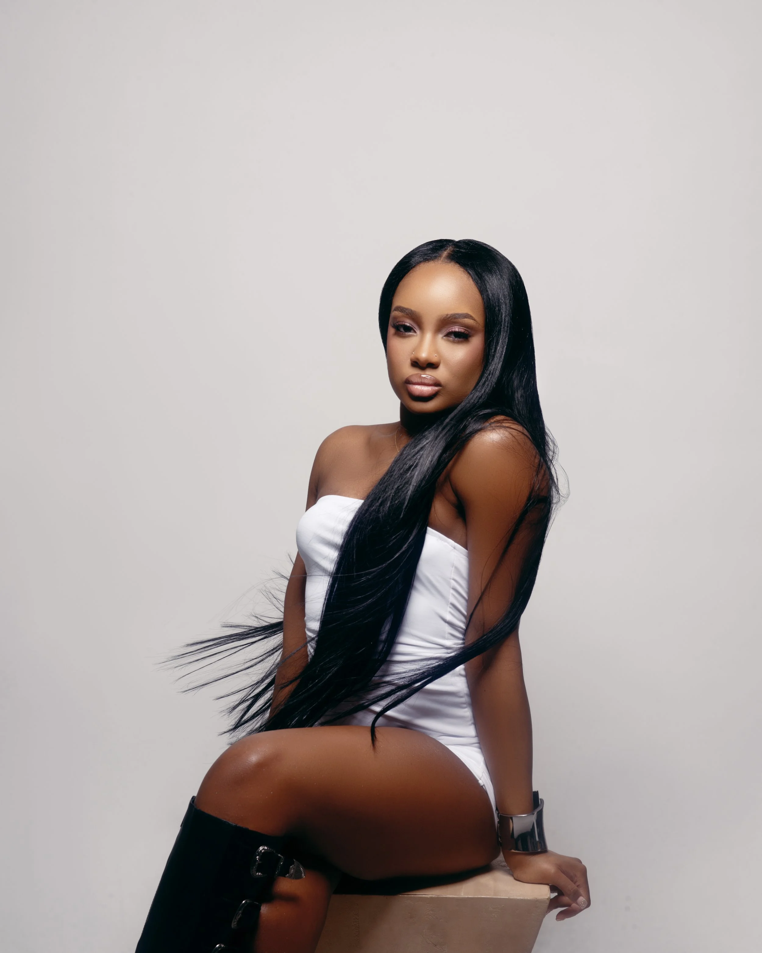 Young woman with long black hair, wearing a white strapless top and black thigh-high boots, sitting on a beige block against a plain gray background.