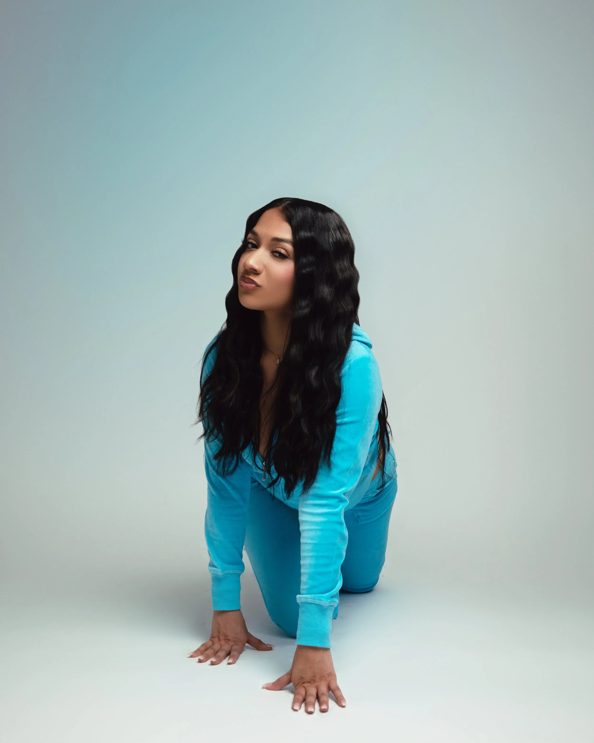 A woman with long black wavy hair in a blue tracksuit on all fours on a plain light background.