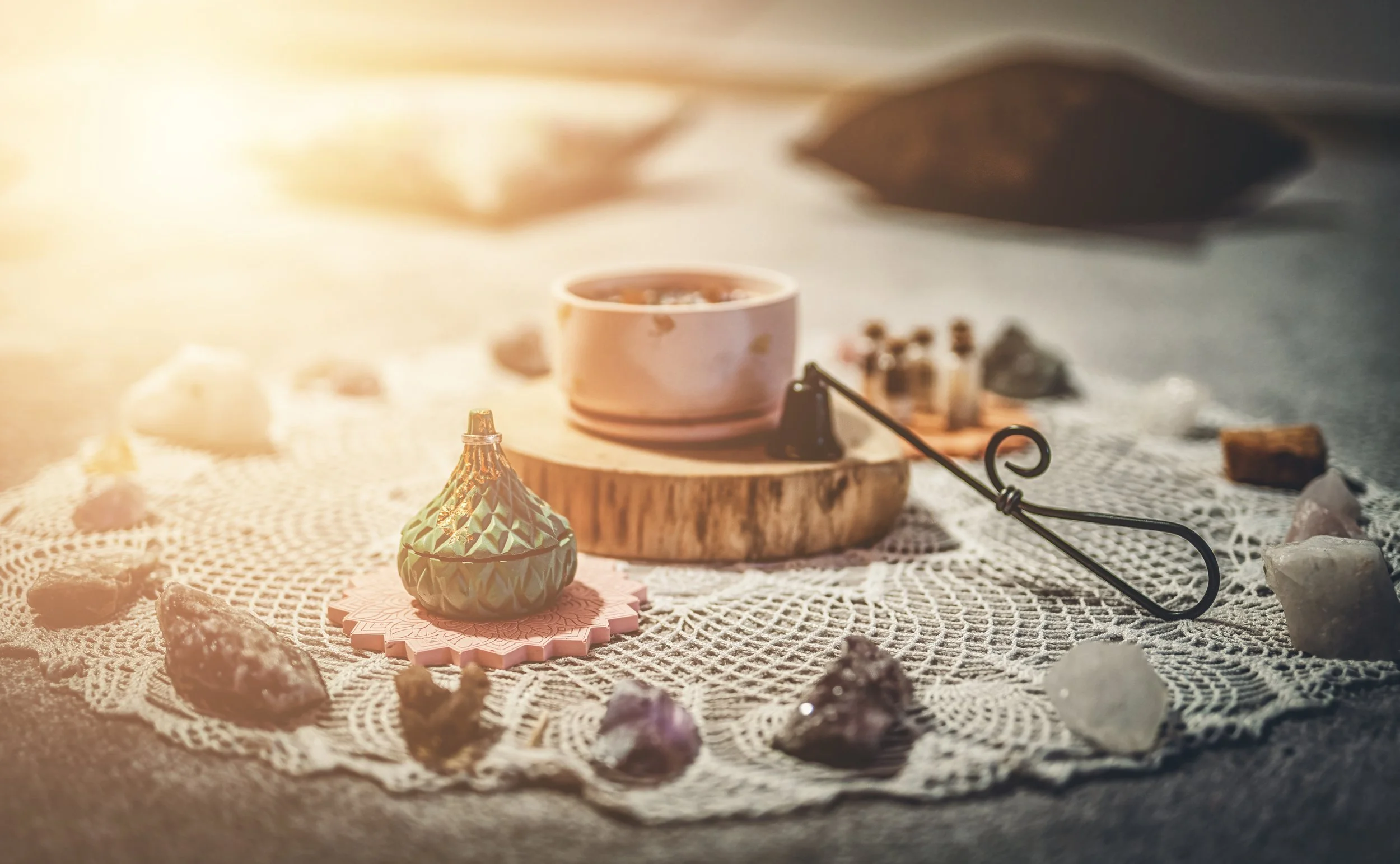 A collection of spiritual or mystical objects arranged on a lace doily, including crystals, a small incense cone, a tea cup, and a candle, with warm sunlight in the background.