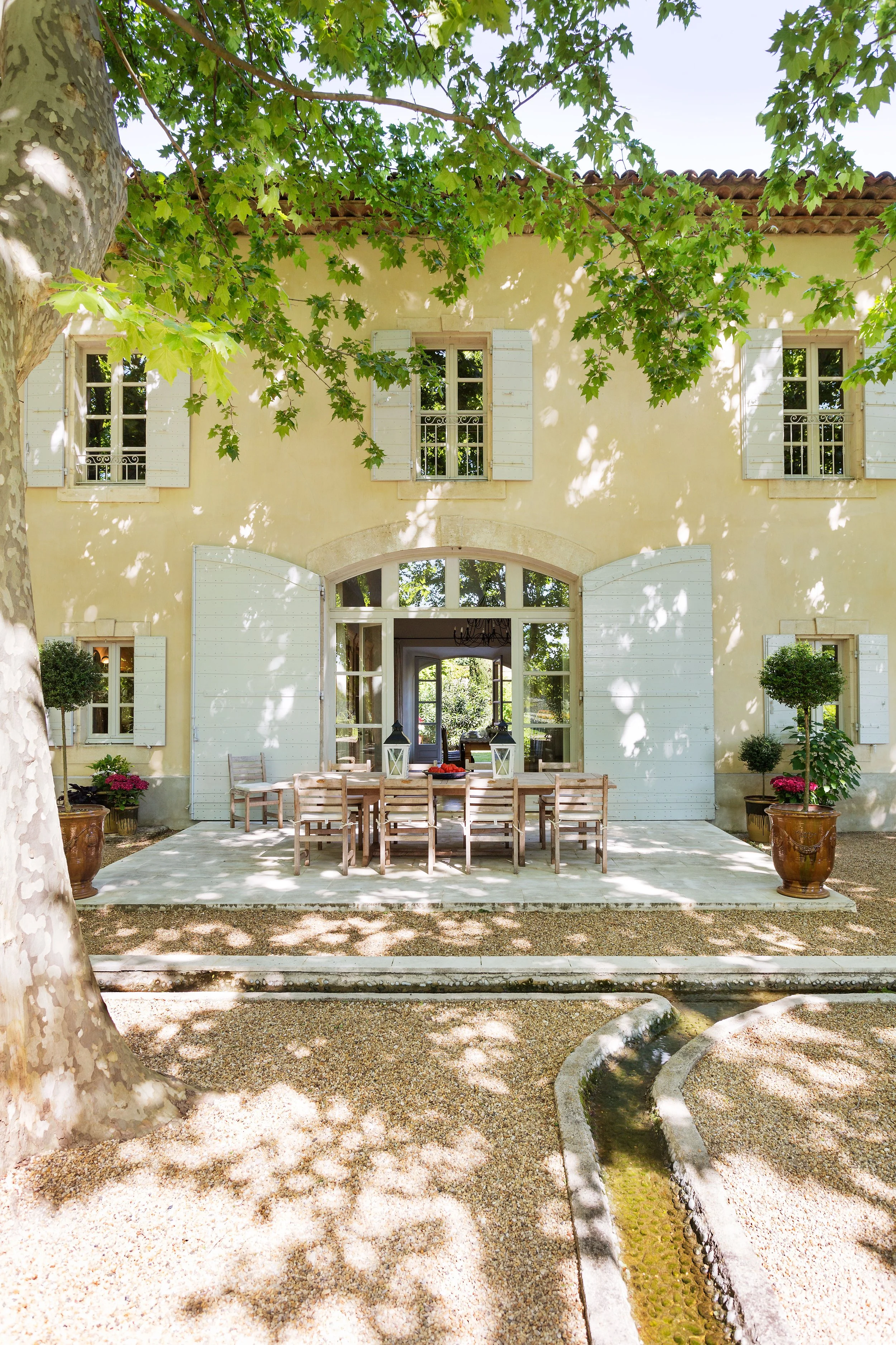 A yellow two-story house with white shutters and a stone arch above large patio doors. An outdoor dining table with chairs is on a tiled patio in front of the house, surrounded by potted plants and trees casting shadows.
