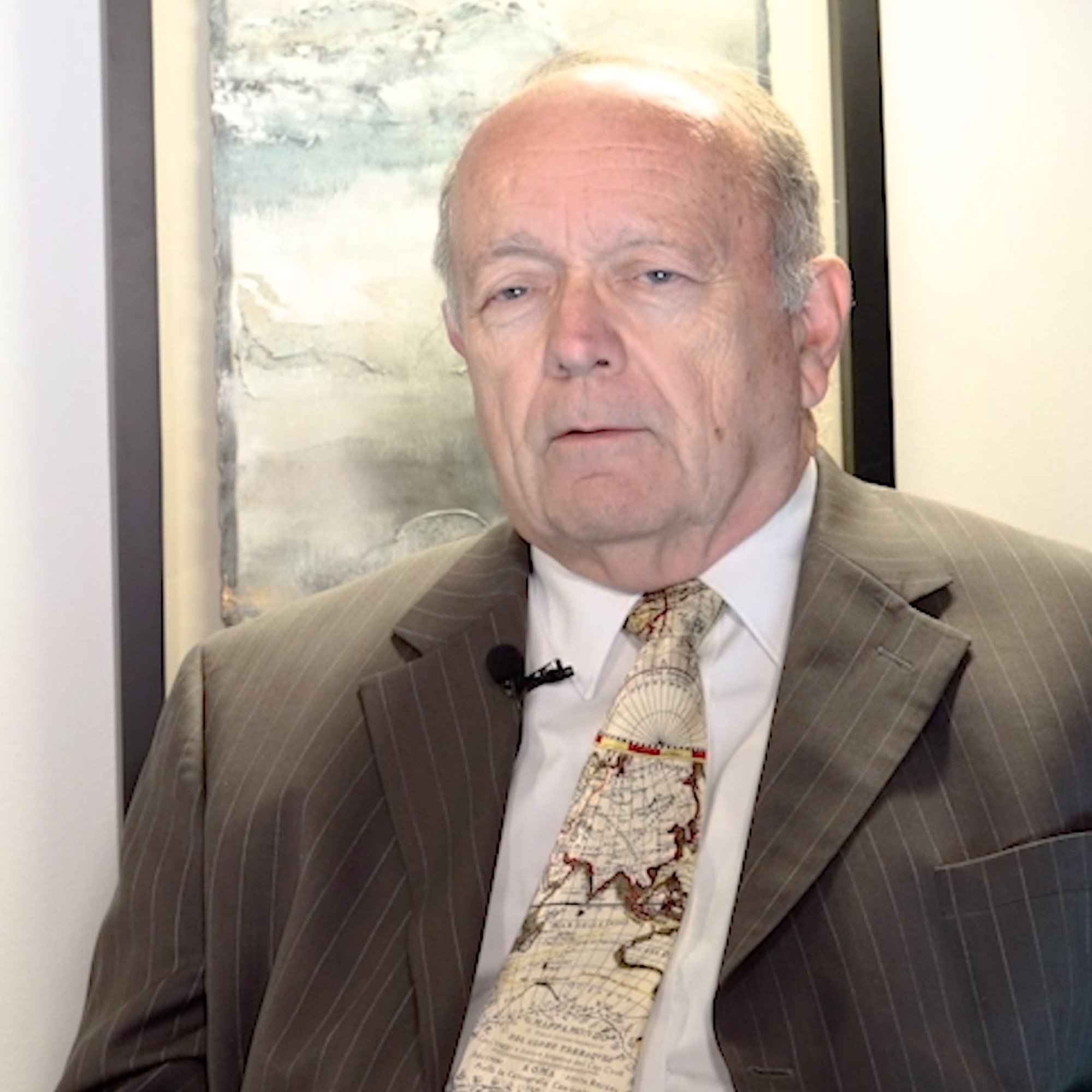 An elderly man with a serious expression, wearing a brown pinstripe suit, a white shirt, and a tie with a map design, sitting in front of a painting with abstract art.