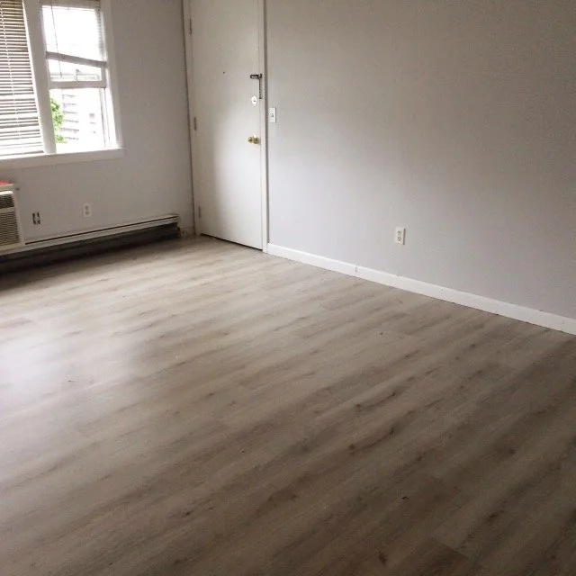 An empty room with wood flooring, white walls, a window with blinds, and a closed door.