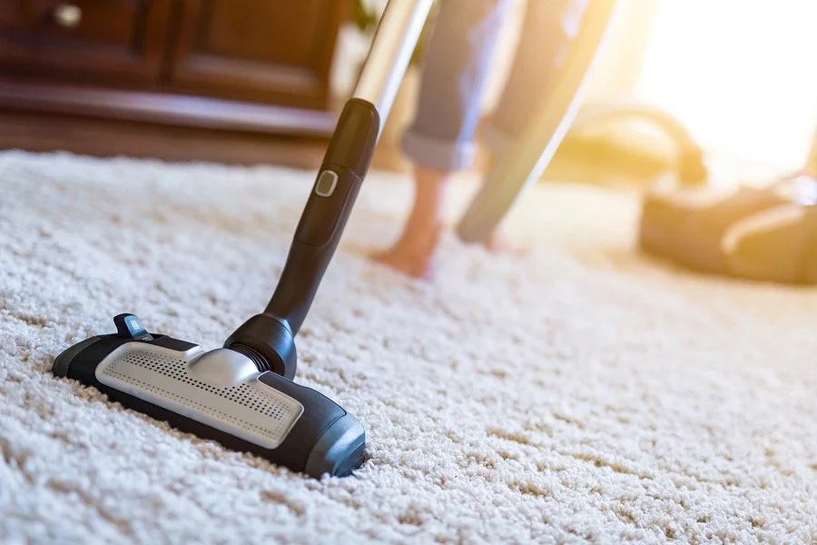 A vacuum cleaner cleaning a light-colored carpet in a living room, with sunlight coming in from a window.