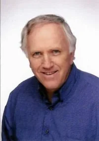 A middle-aged man with gray hair wearing a blue button-up shirt, smiling against a plain white background.