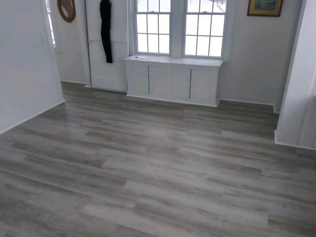 Empty room with light-colored wood flooring, white walls, a white bench under a window, a door with hanging clothing, and a framed picture on the wall.