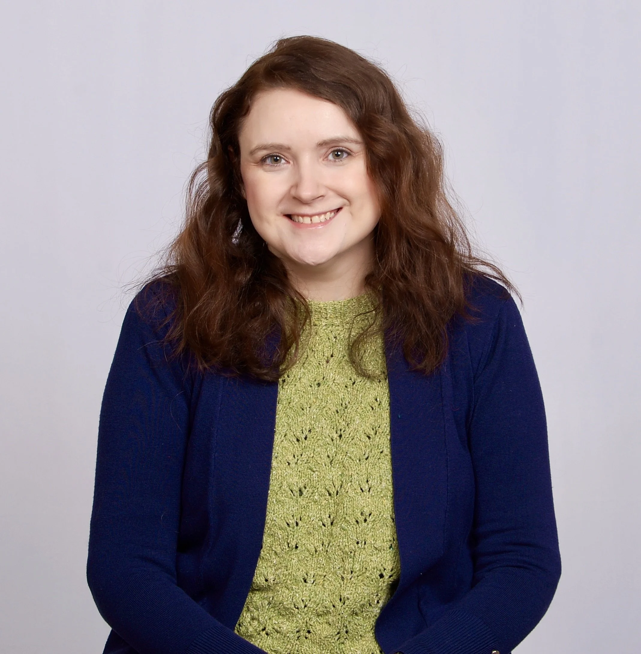 A woman with long, wavy, auburn hair and blue eyes, smiling and wearing a navy blue jacket over a green textured top, standing against a plain light gray background.