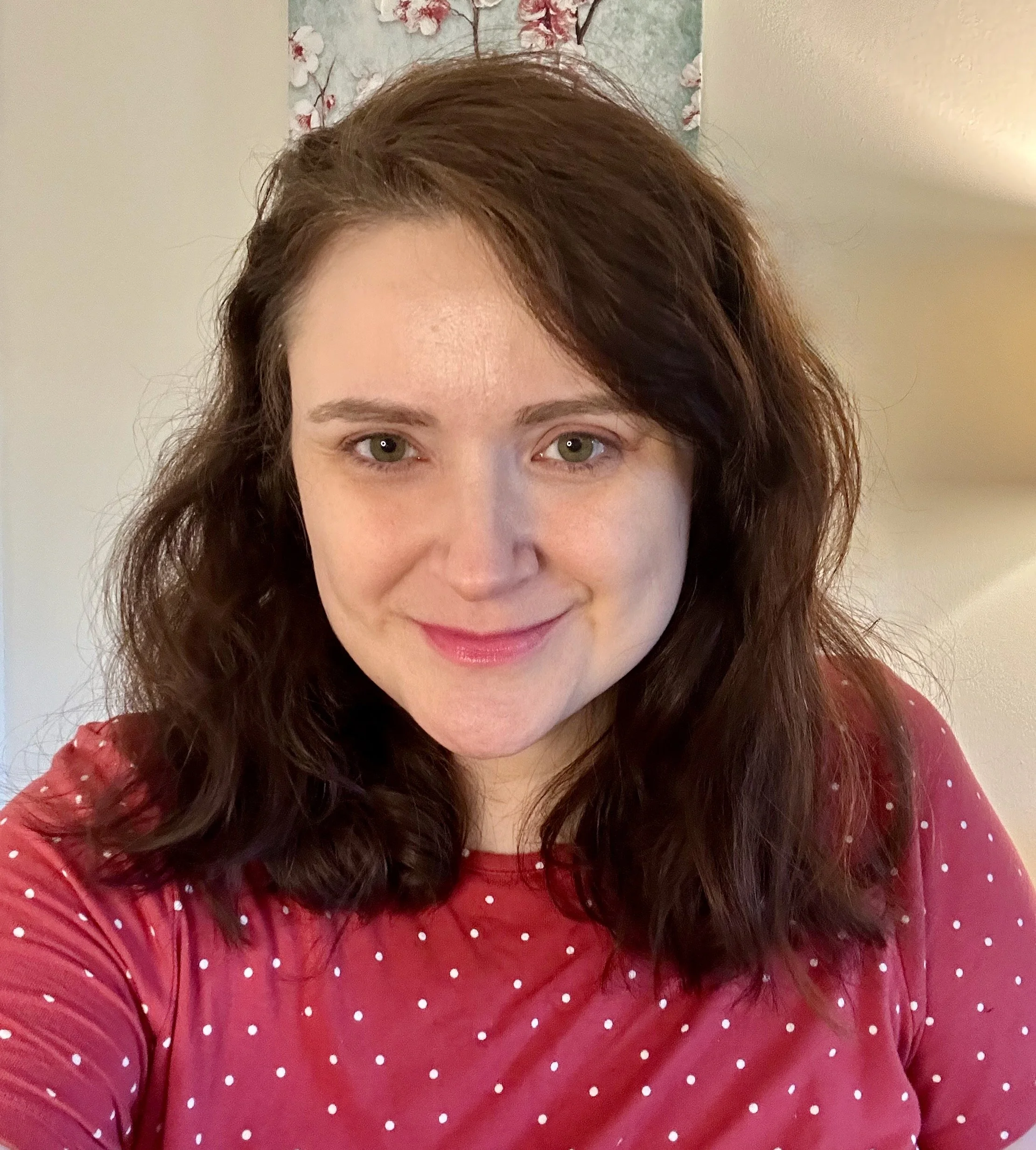 Close-up of a woman with curly brown hair, green eyes, and a pink shirt with white polka dots, smiling softly with a partially visible floral painting in the background.