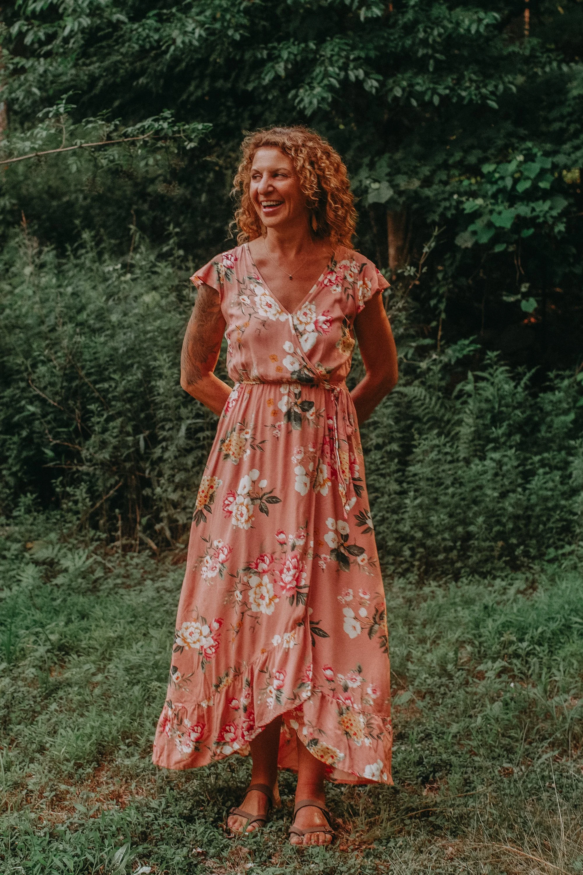 A woman in a pink floral maxi dress standing outdoors in a grassy area with greenery behind her, smiling and looking to her left.
