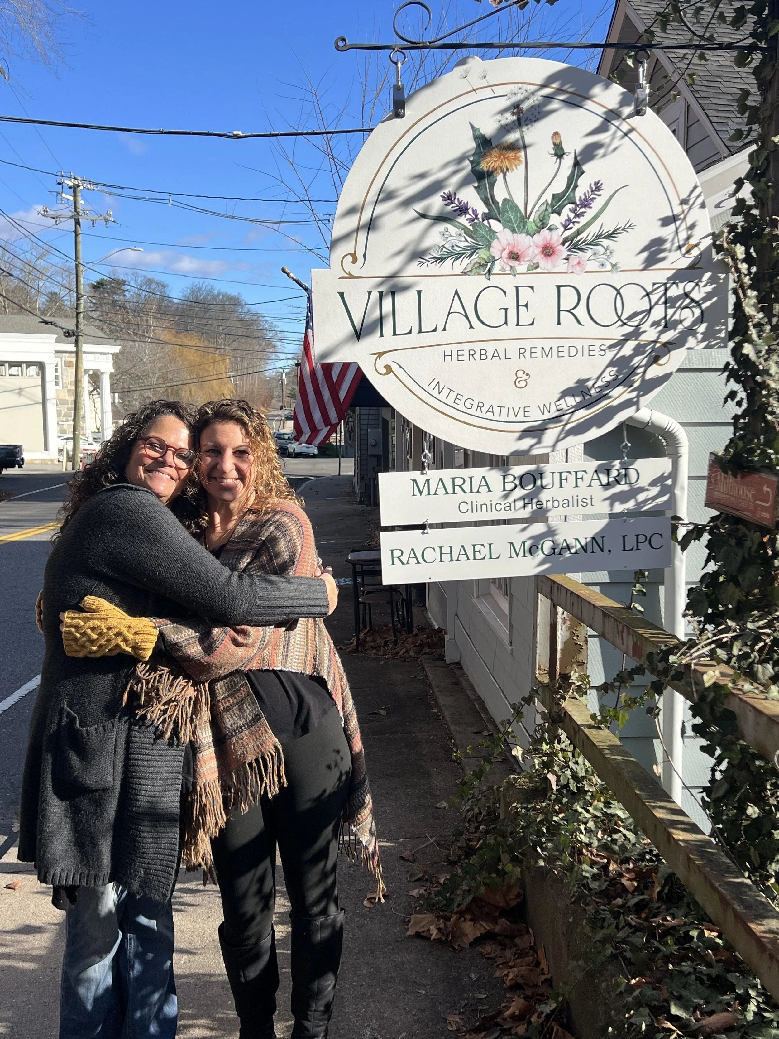 Two women hugging and smiling on a sidewalk beside a sign for Village Roots Herbal Remedies and Wellness, with a clear blue sky and trees in the background.