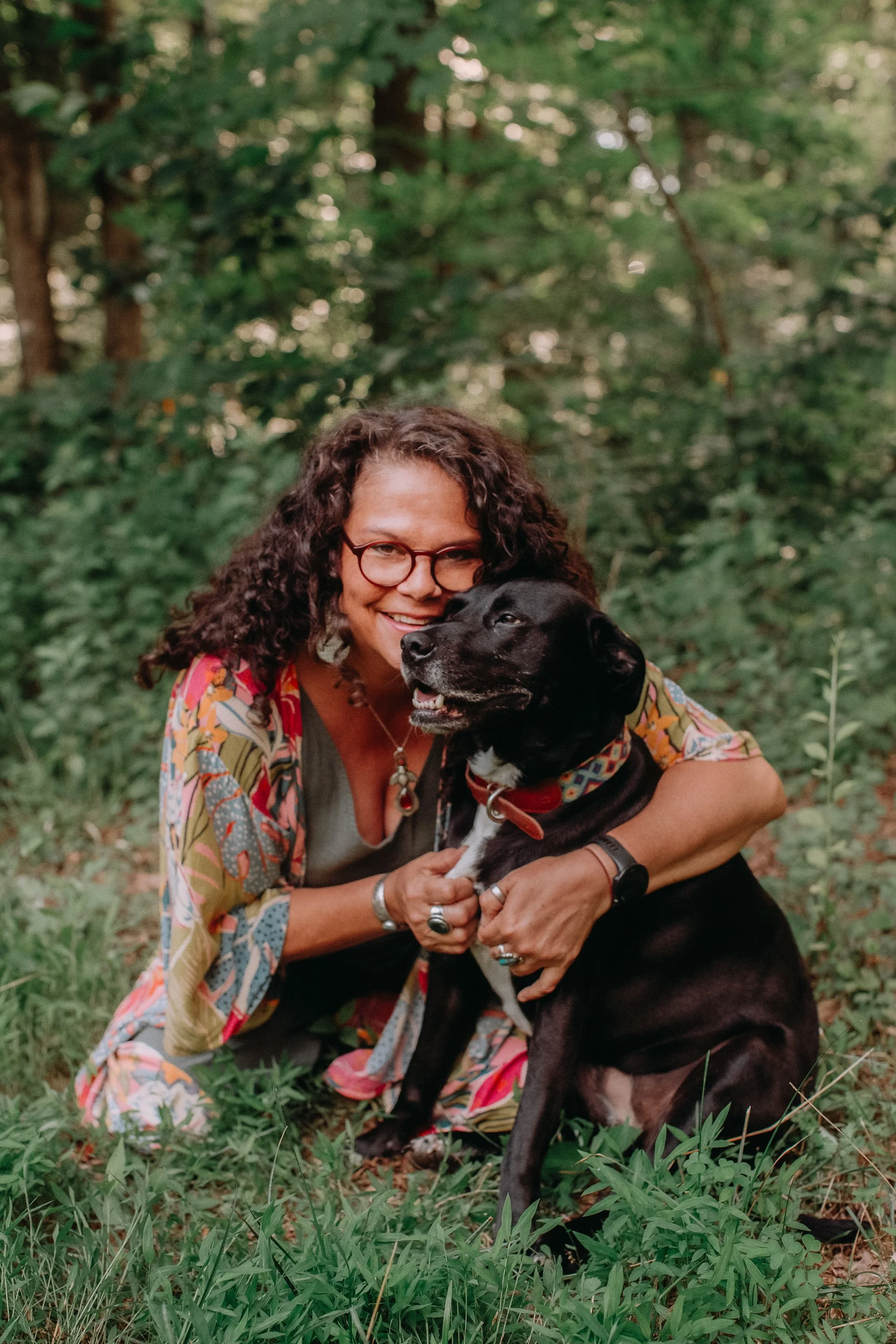 A woman with glasses and curly hair smiling and hugging a black dog in a forest setting.