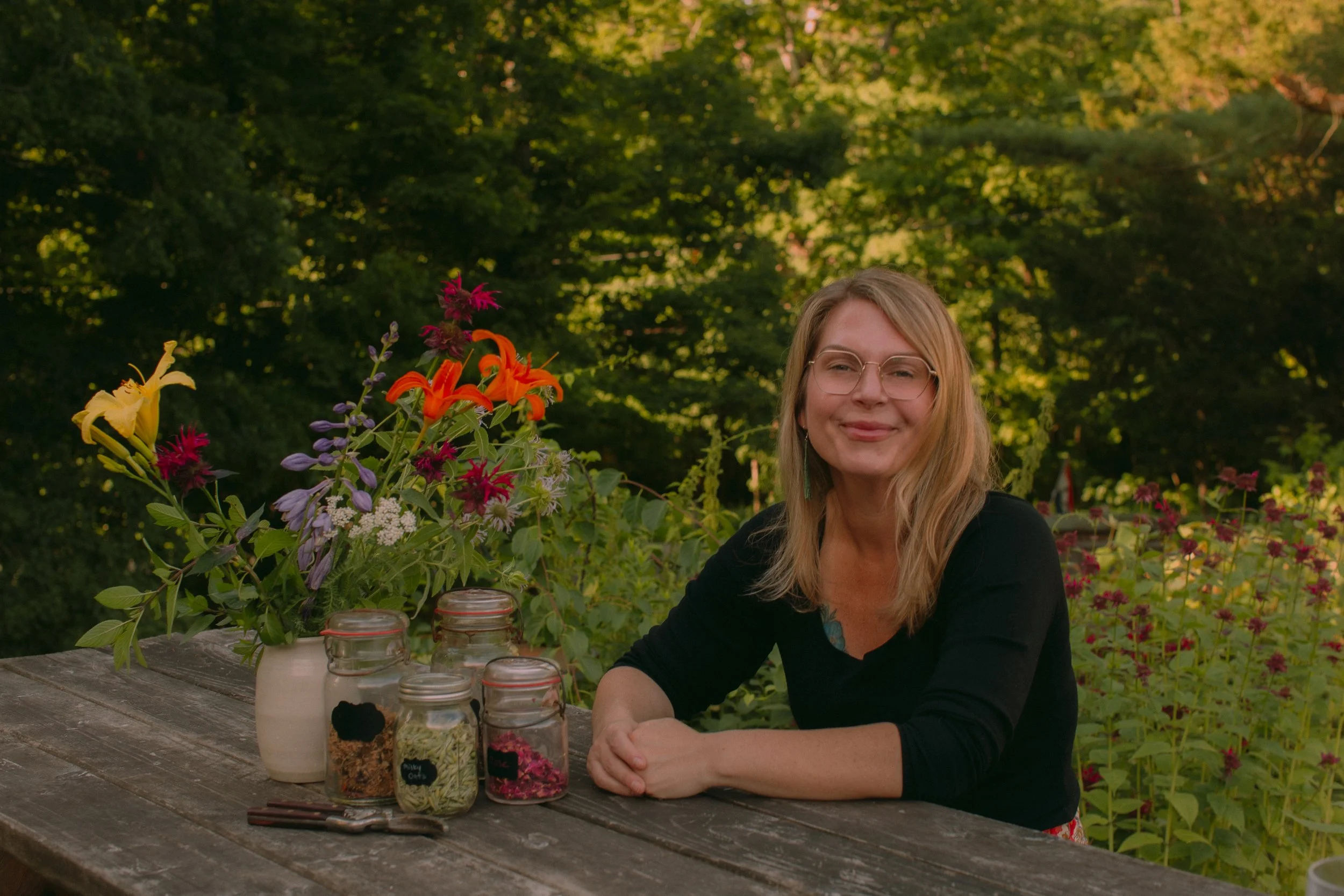 A woman sitting at a wooden table outdoors, with a large bouquet of colorful flowers, jars with herbs and flowers, and some utensils, during daylight with a green leafy background.