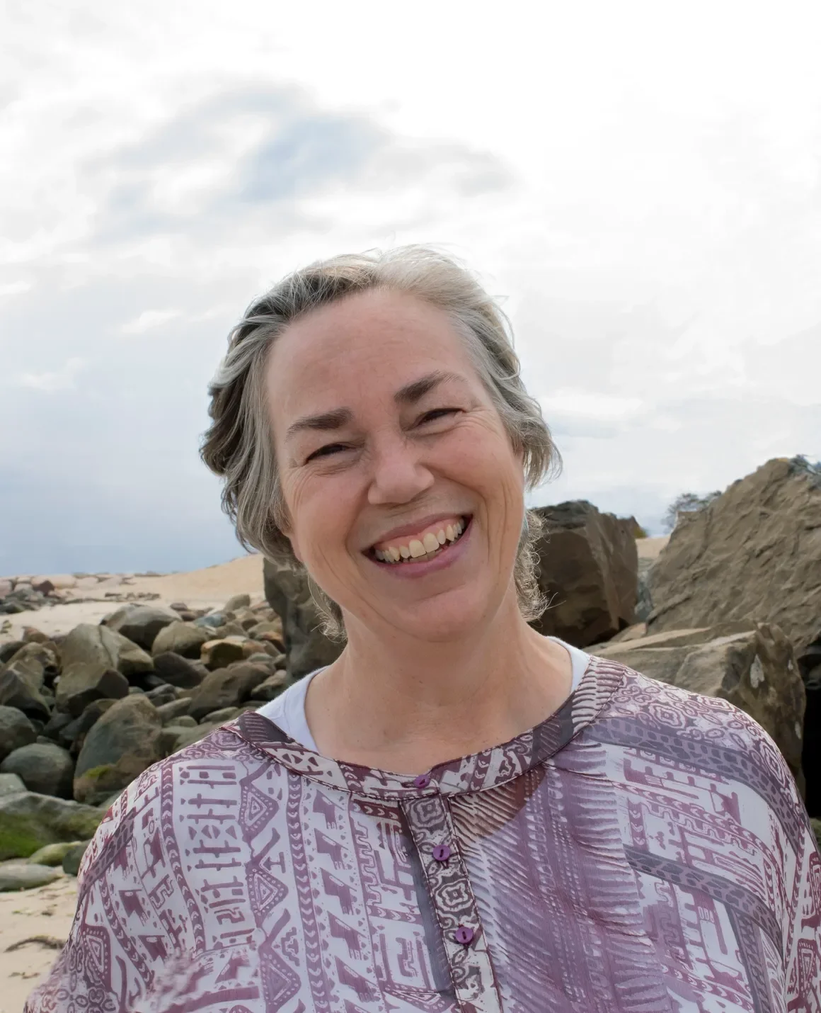 A smiling older woman with short gray hair standing on a rocky beach with large stones and sand, under a cloudy sky.