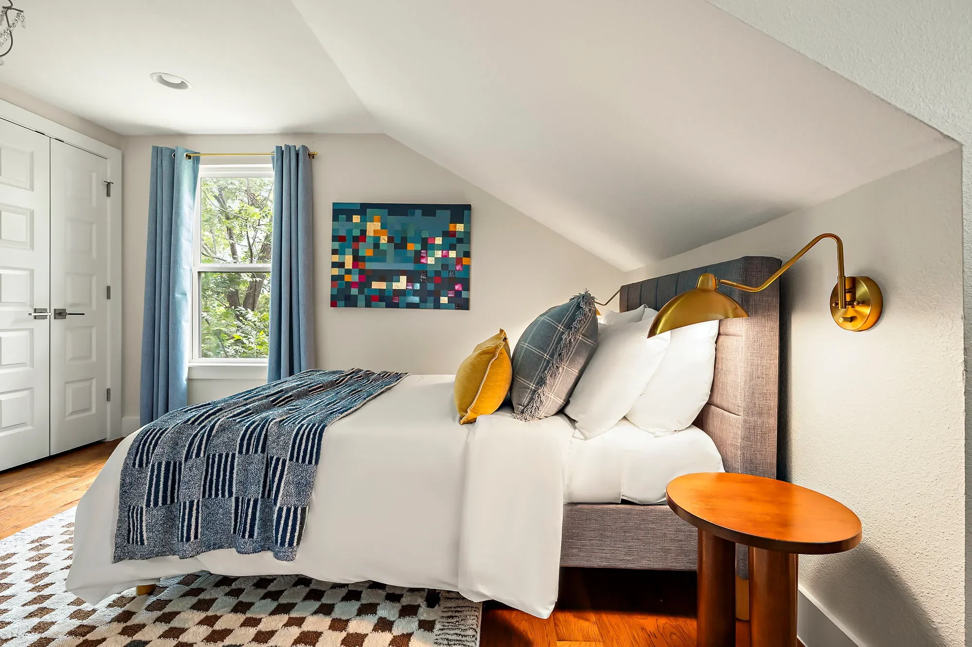 Mid-century modern bedroom with geometric and checkered wall art and rug, gold wall sconces, wood nightstand, and blue floor-to-ceiling curtains.