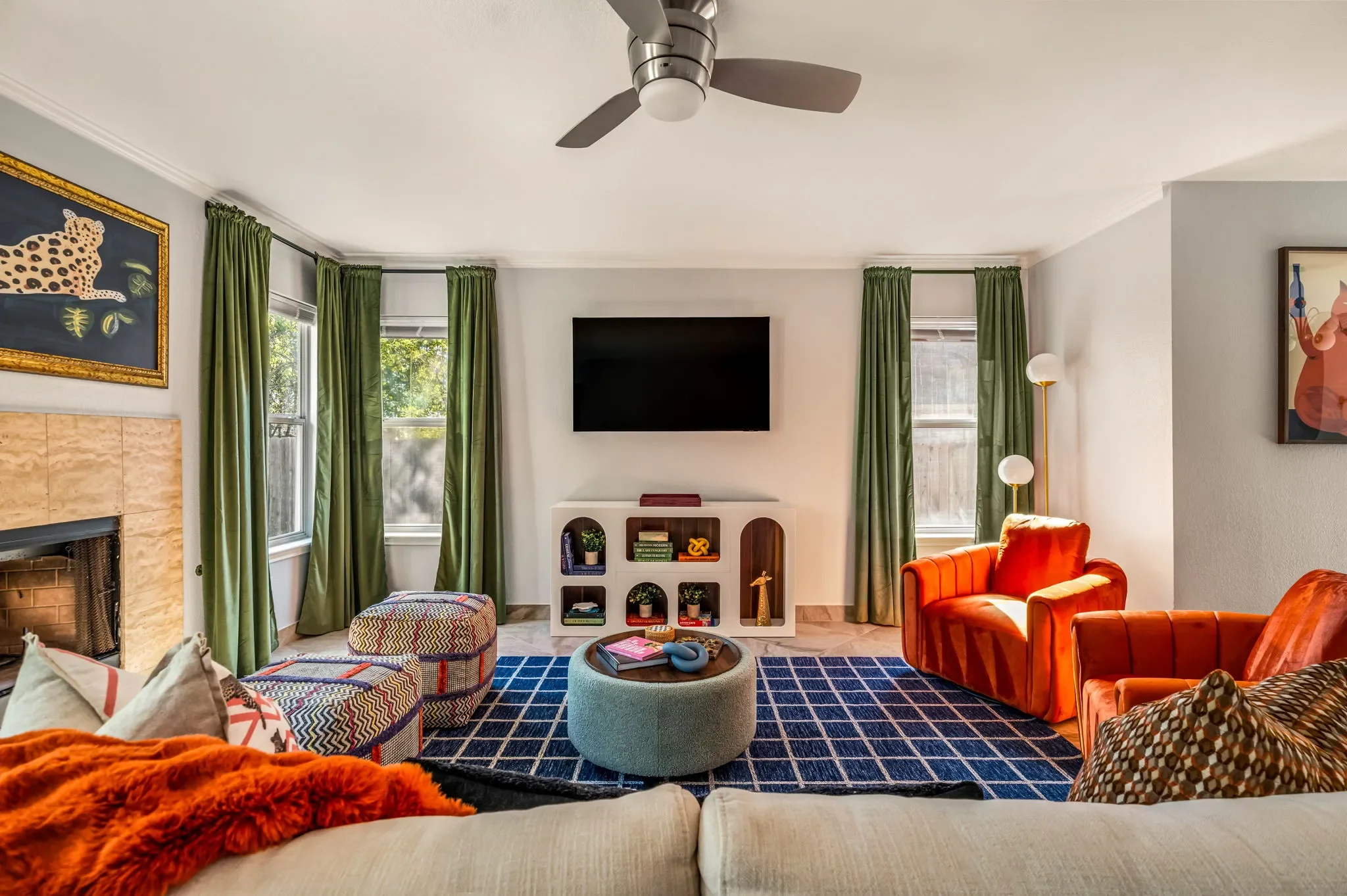 Colorful living room featuring a blue checkered area rug, green floor-to-ceiling curtains, orange accent chairs, and a wall-mounted TV.