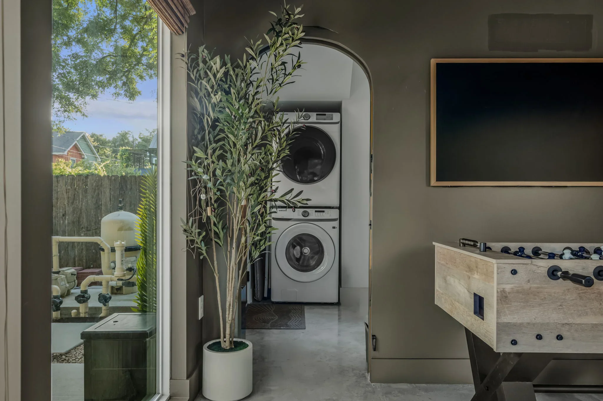Airbnb game room design with stacked washer dryer in arched nook, TV, olive tree, and foosball table — Austin TX.