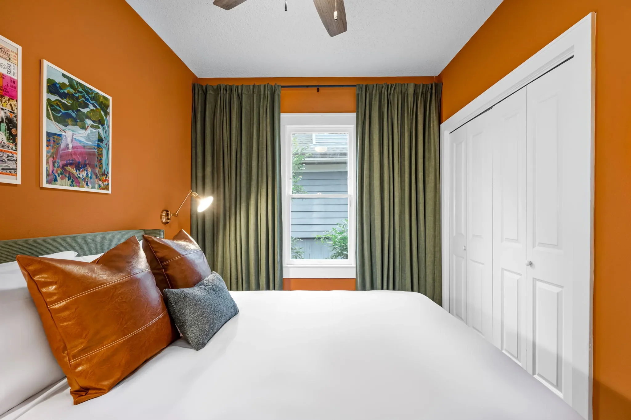 Short-term rental bedroom design with bold orange walls, green curtains, leather pillow, and colorful travel wall art — Austin TX.