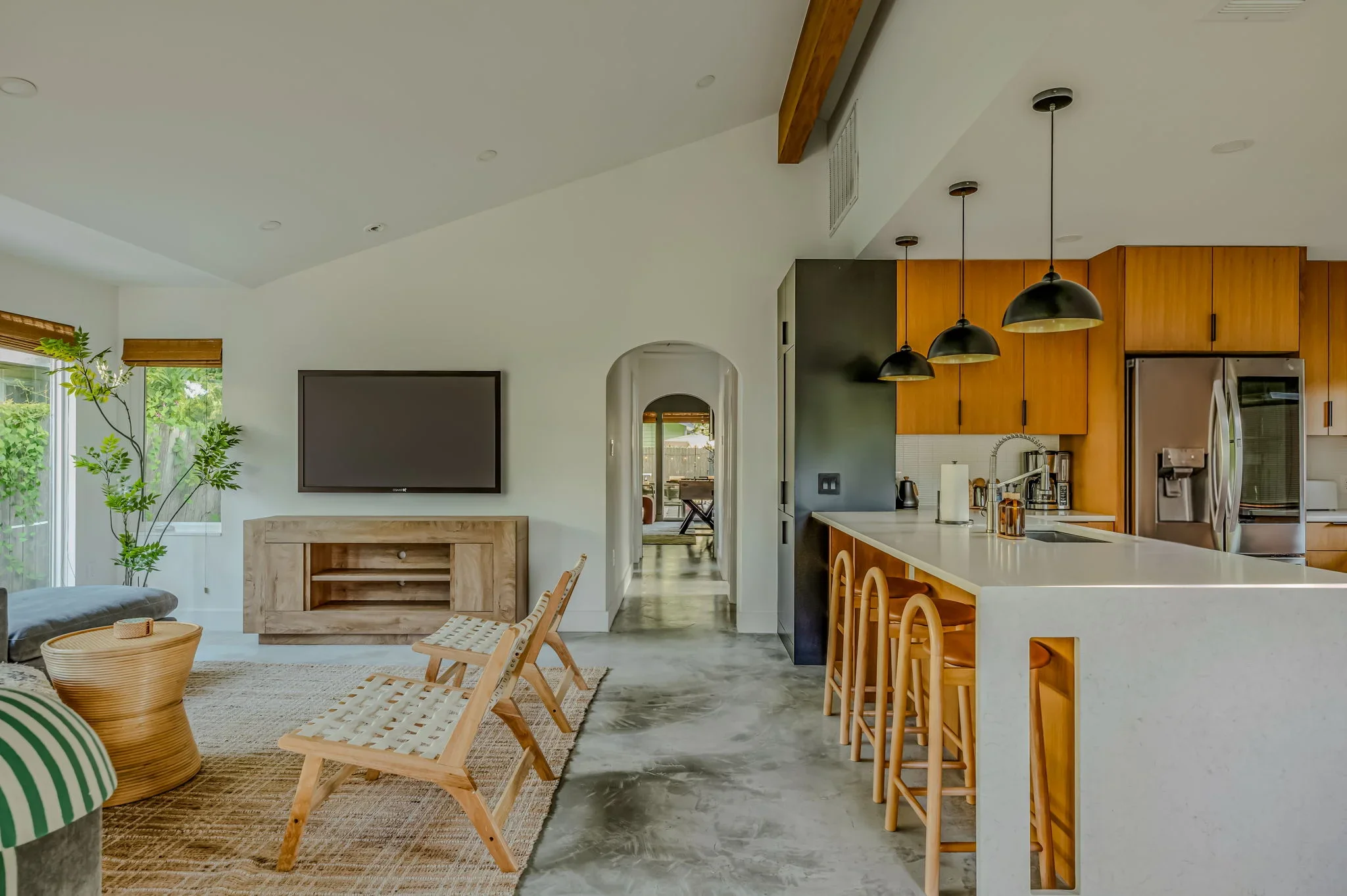 Airbnb open plan design with wood island bar stools, arched doorway, TV, warm wood kitchen cabinetry, and woven accent chairs — Austin TX.