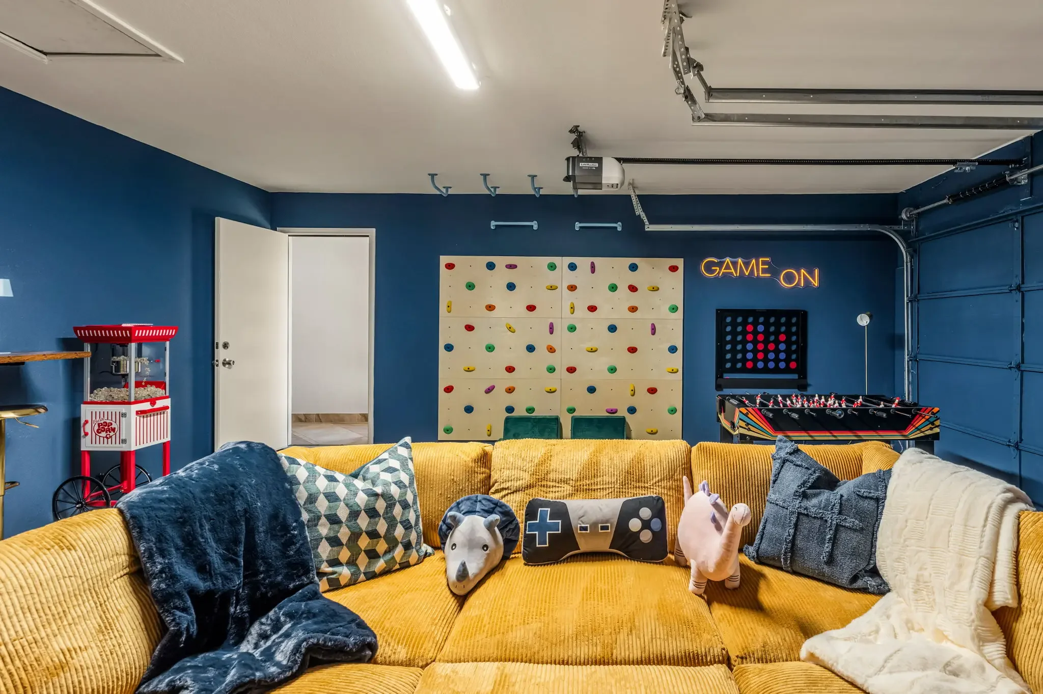 Short-term rental garage game room with blue accent walls, yellow sectional, rock climbing wall, and Game On neon sign — Austin TX.