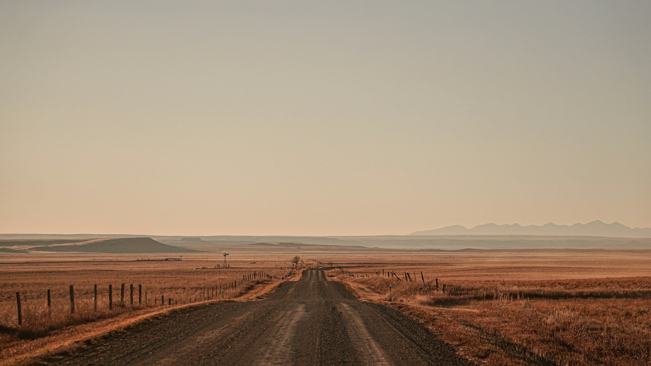 Dirt country road stretching into the distance through flat, open grassland with fences on both sides and hills on the horizon under a clear sky.