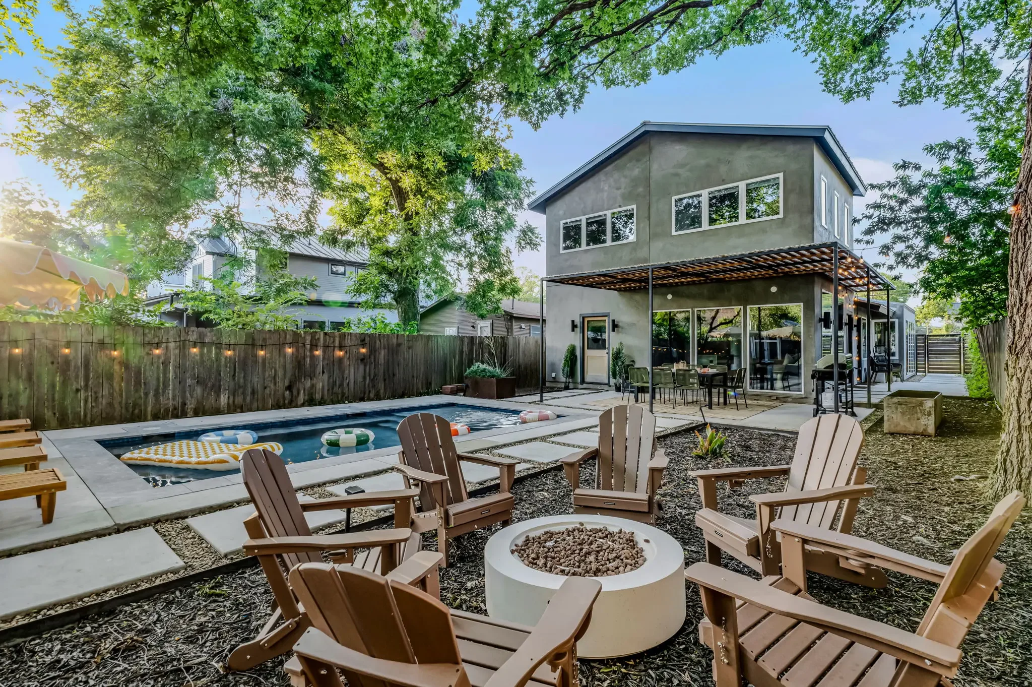 Airbnb backyard design with fire pit, wood Adirondack chairs, lap pool, pergola dining area, and mature oak trees — Austin TX.