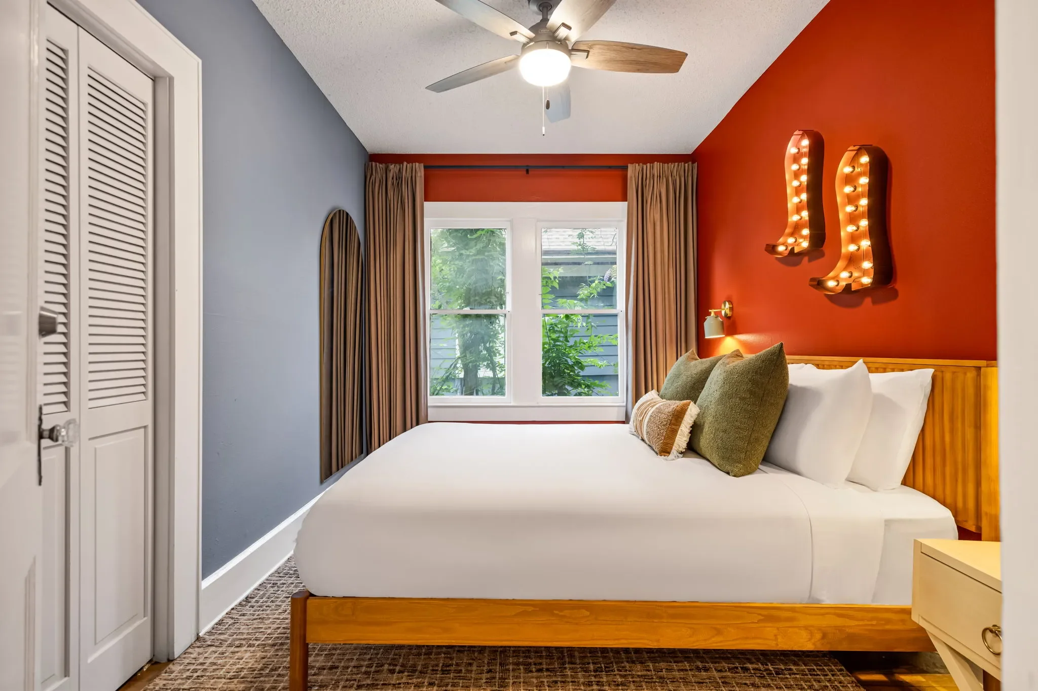 Short-term rental bedroom design with orange accent wall, blue-gray walls, wood bed frame, green pillows, and cowboy boot marquee lights — Austin TX.