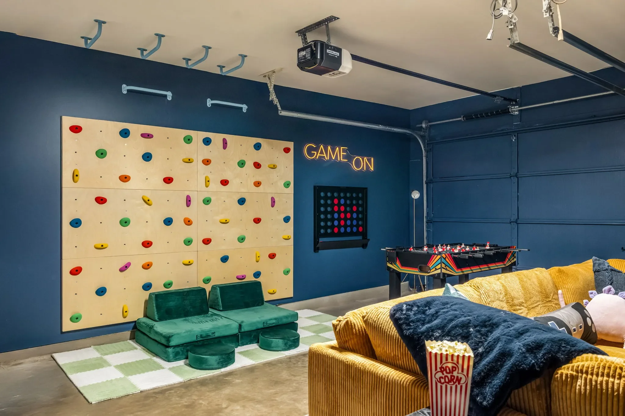 Converted garage game room with navy blue walls, a “Game On” neon sign, indoor residential rock climbing wall, and game tables.