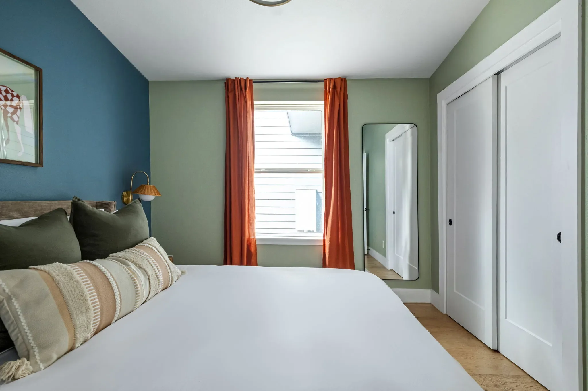 Short-term rental bedroom design with blue and sage green accent walls, wood headboard, orange curtains, and leaning mirror — Austin TX.