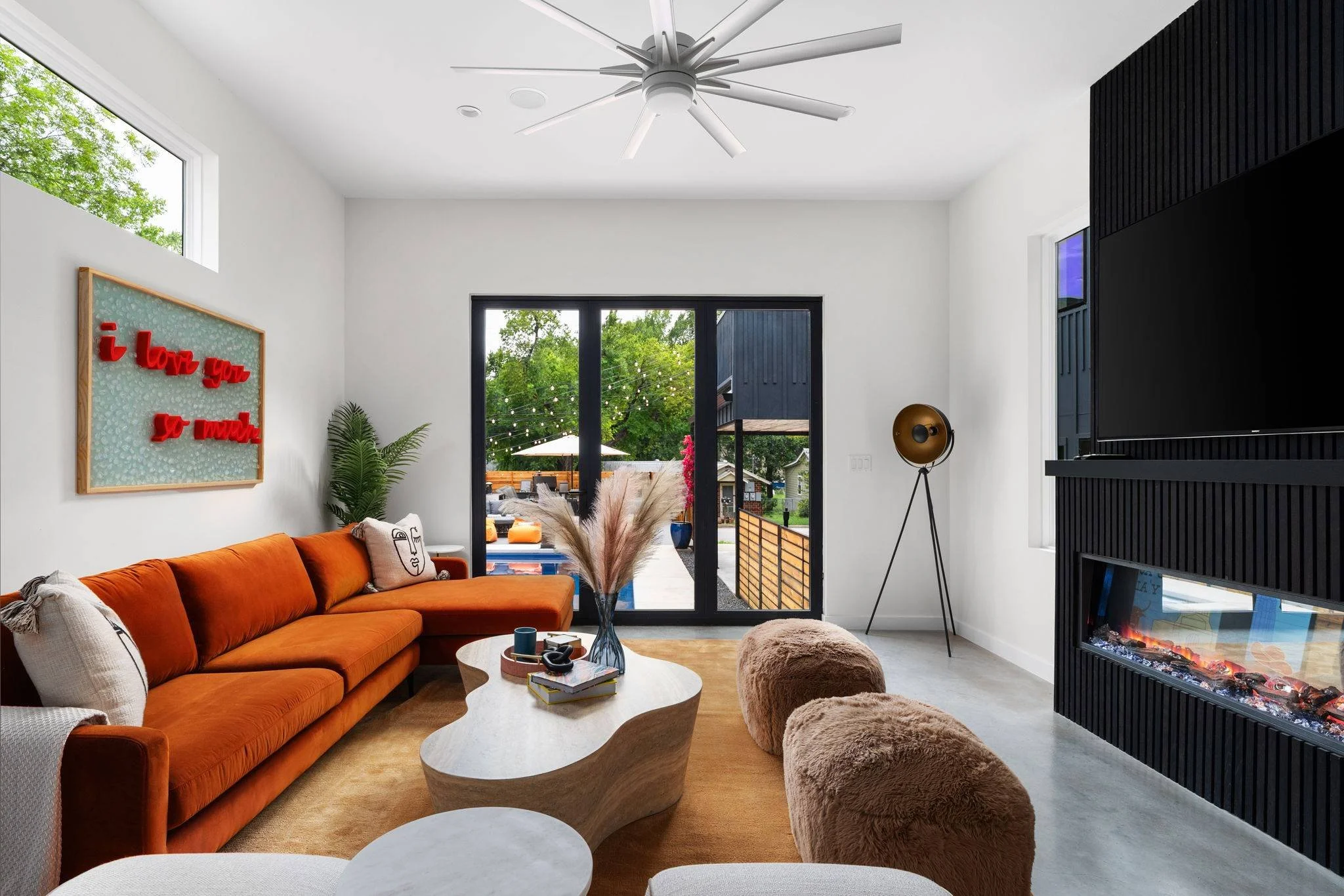 Modern living room in an Austin short-term rental featuring an orange sectional sofa, a travertine coffee table, fur poufs, a black fireplace wall, and sliding glass doors opening to the backyard.