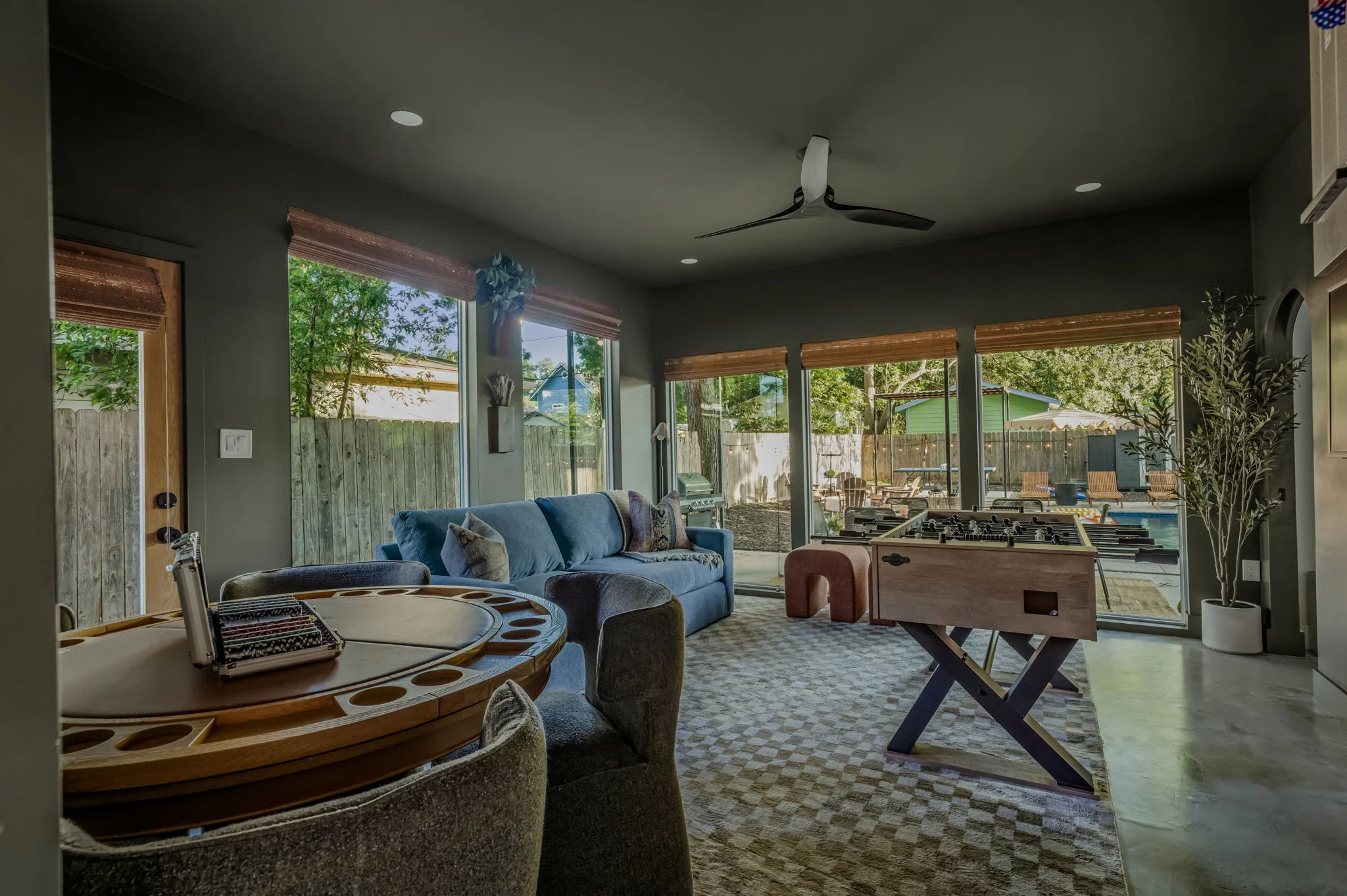 Airbnb game room design with round poker table, blue sofa, foosball table, checkered rug, and sliding glass doors to pool — Austin TX.