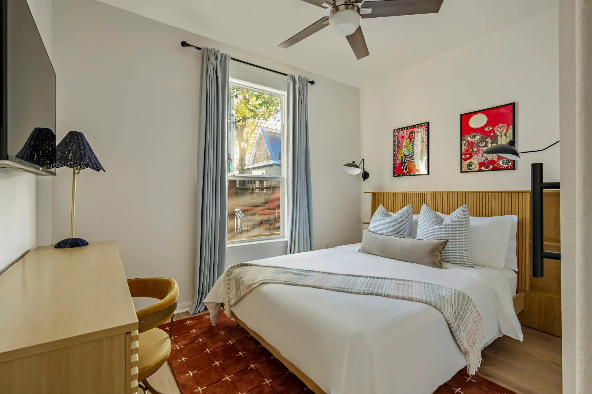 Bedroom with eclectic psychedelic wall art, wooden bed frame with built-in nightstands, light blue floor-to-ceiling curtains, a rust area rug, and a desk area.