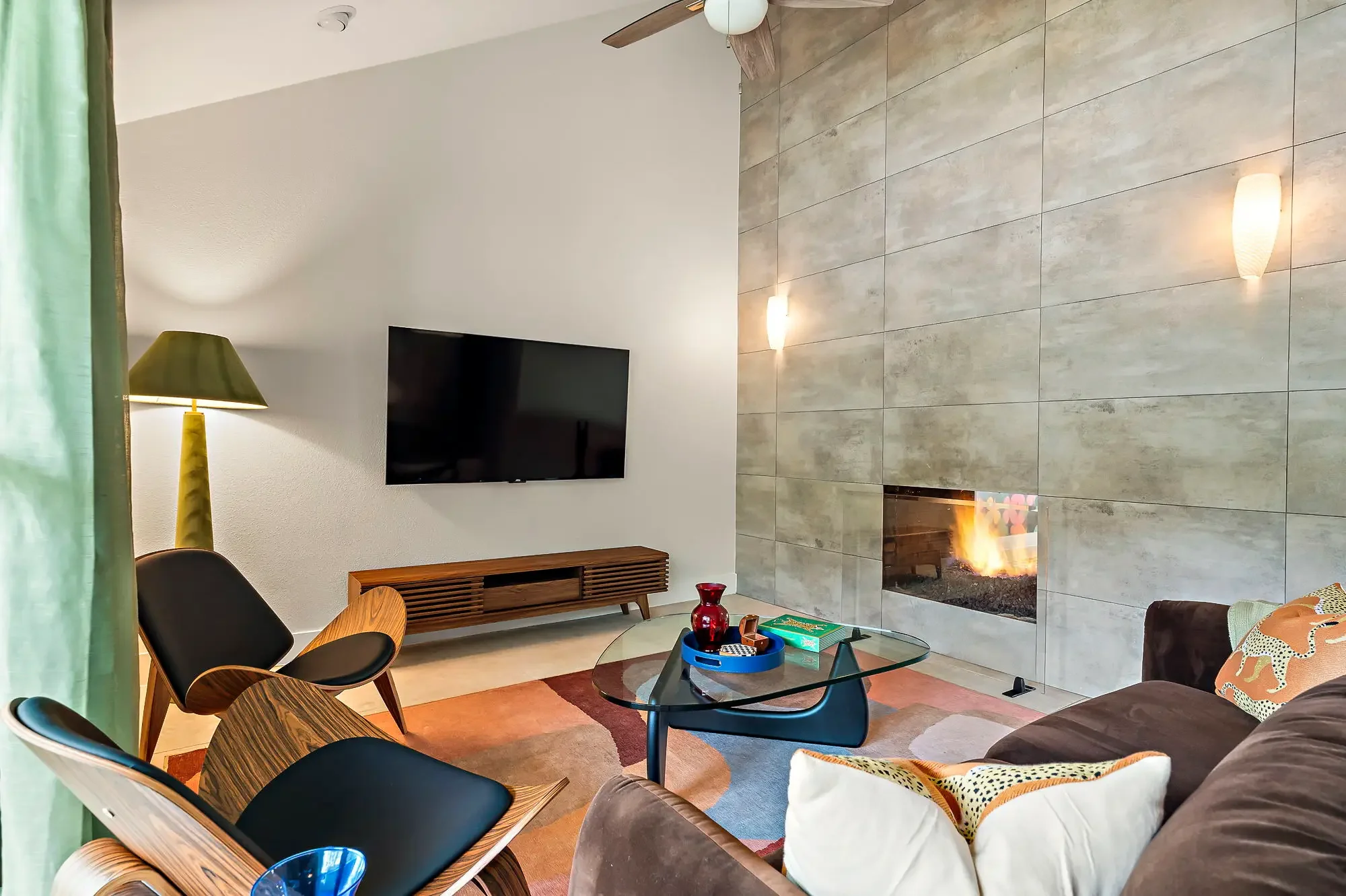 Short-term rental sitting room design with concrete fireplace wall, mid-century chairs, glass coffee table, and green lamp — Austin TX.