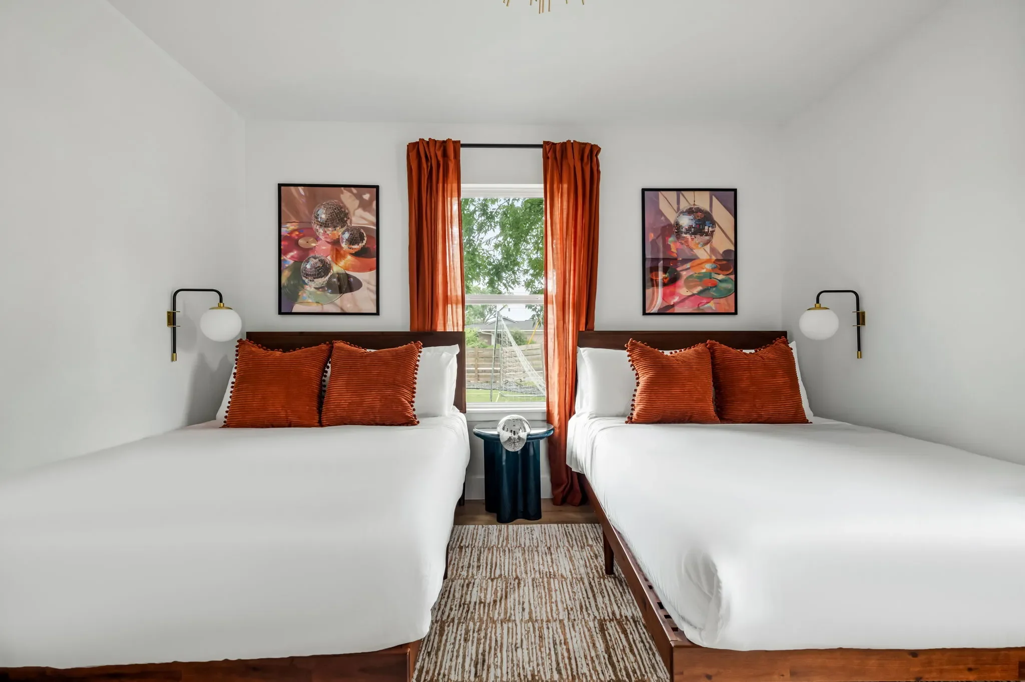 Short-term rental double bedroom design with two queen beds, rust orange pillows, wood headboards, wall sconces, and colorful art — Austin TX.