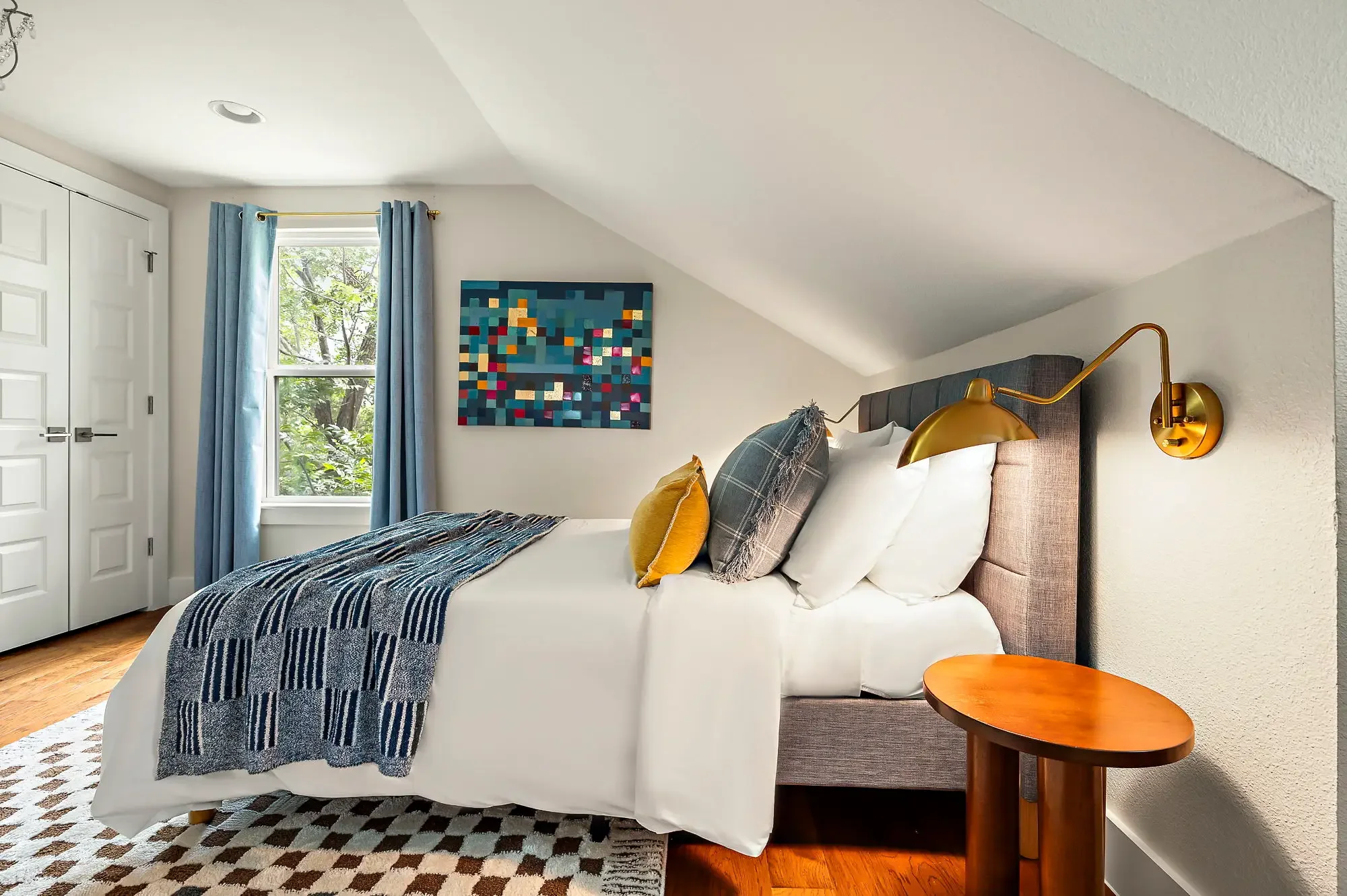 Airbnb attic bedroom design detail with gray upholstered headboard, gold sconce, yellow pillows, blue throw, and mosaic wall art — Austin TX.