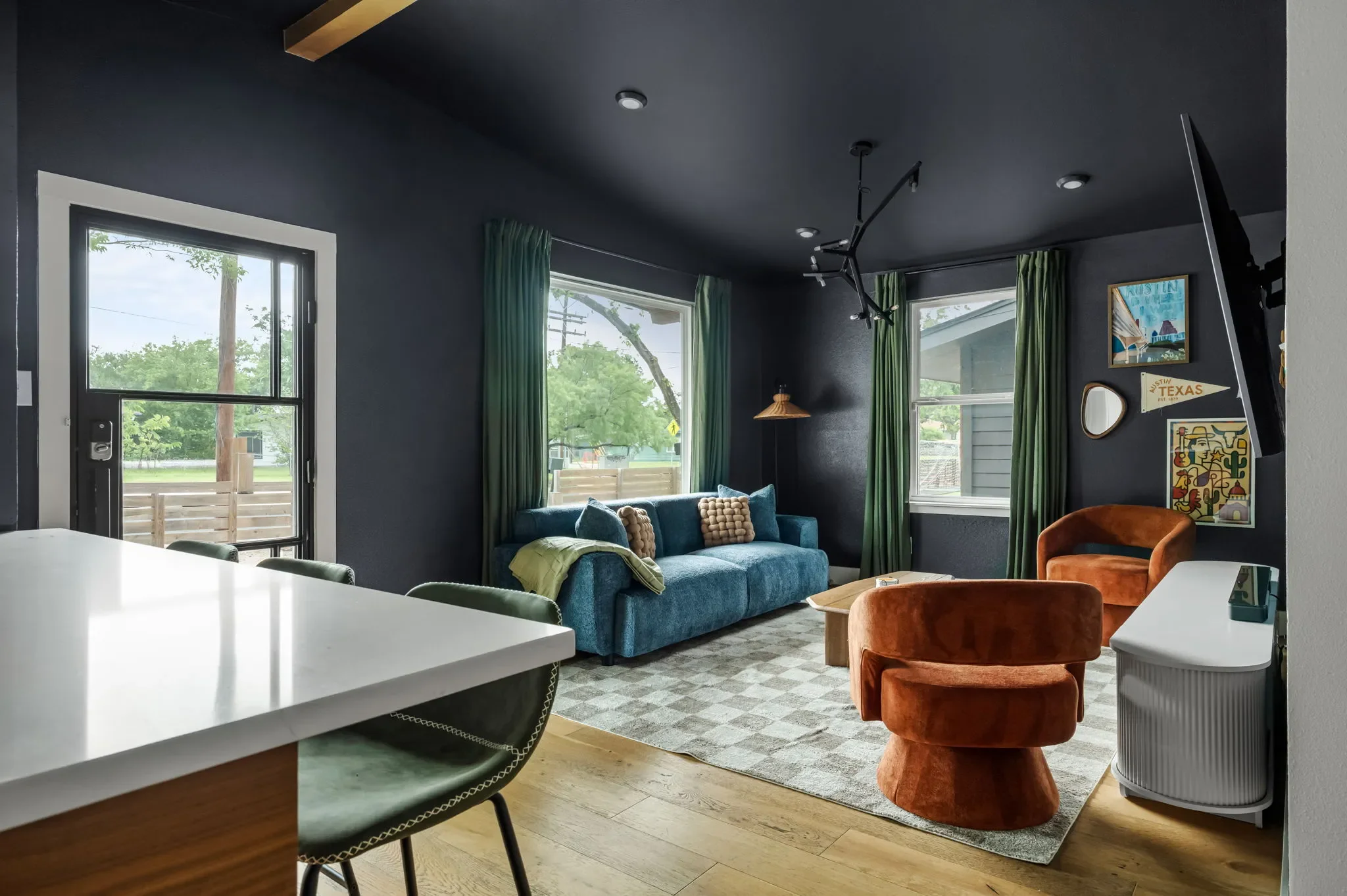 Short-term rental open plan design with dark walls, blue sofa, orange accent chairs, checkered rug, and green curtains — Austin TX.